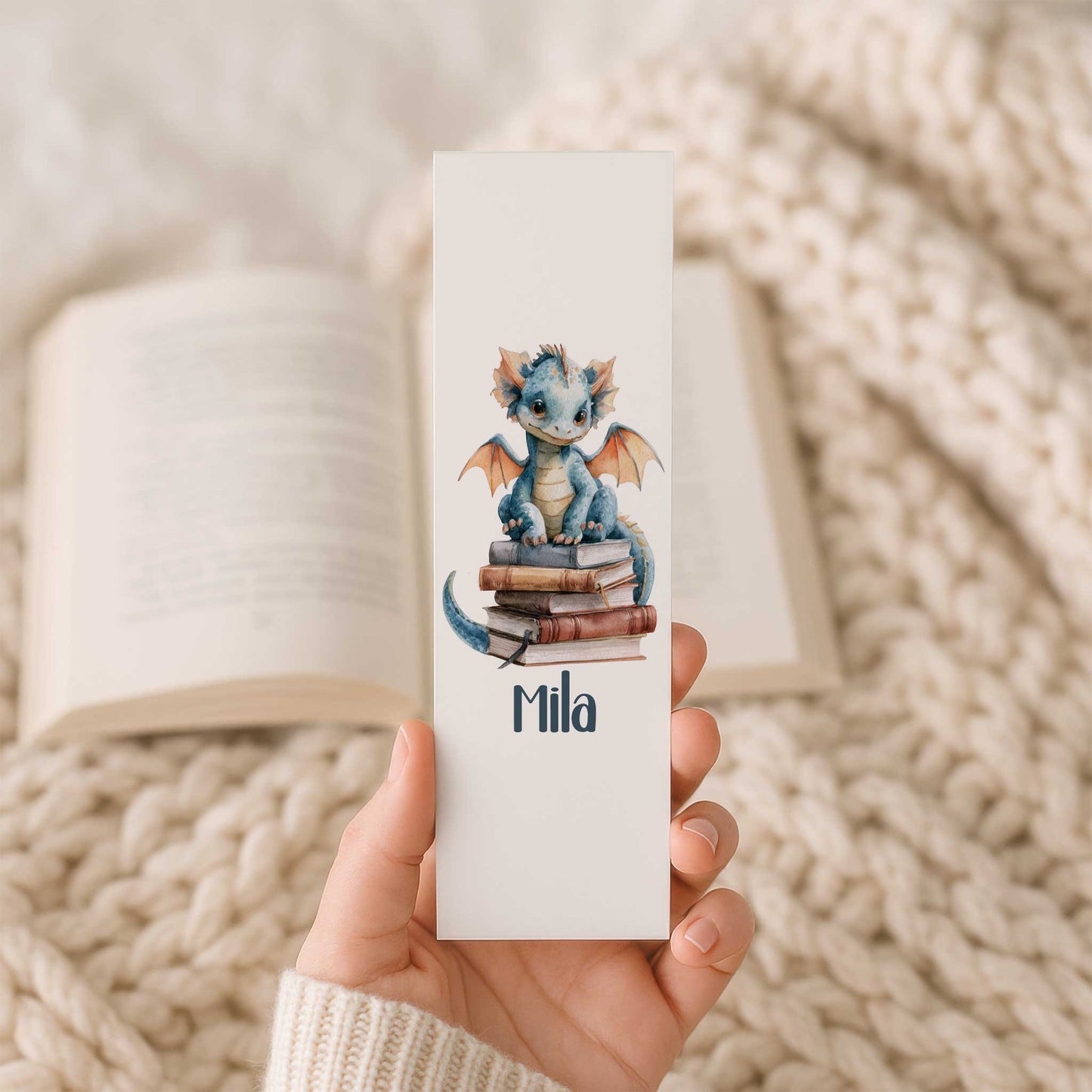 Hand houdt een boekenlegger vast met een blauw draakje op een stapel boeken en de naam Mila voor een open boek.