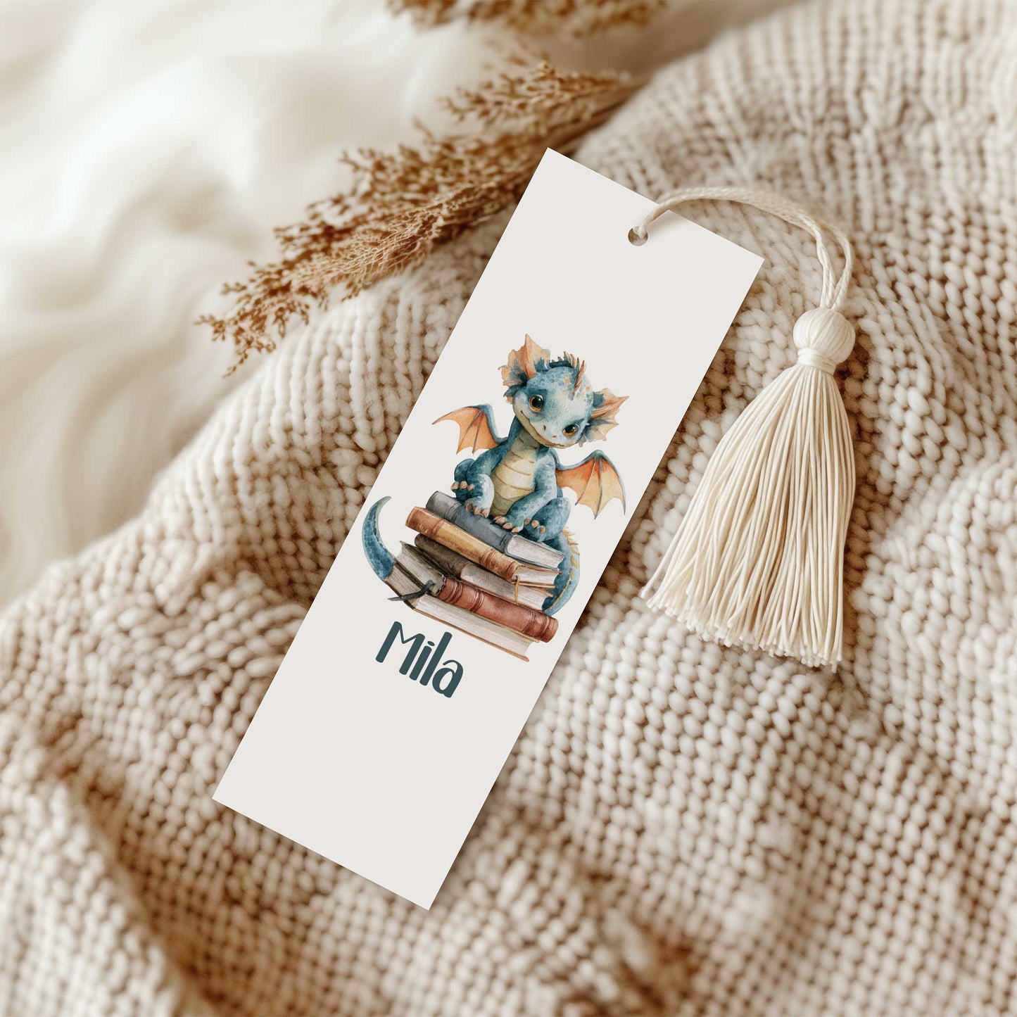 Boekenlegger met een blauw draakje op een stapel boeken en de naam Mila op een gebreide beige deken naast een crèmekleurig kwastje.