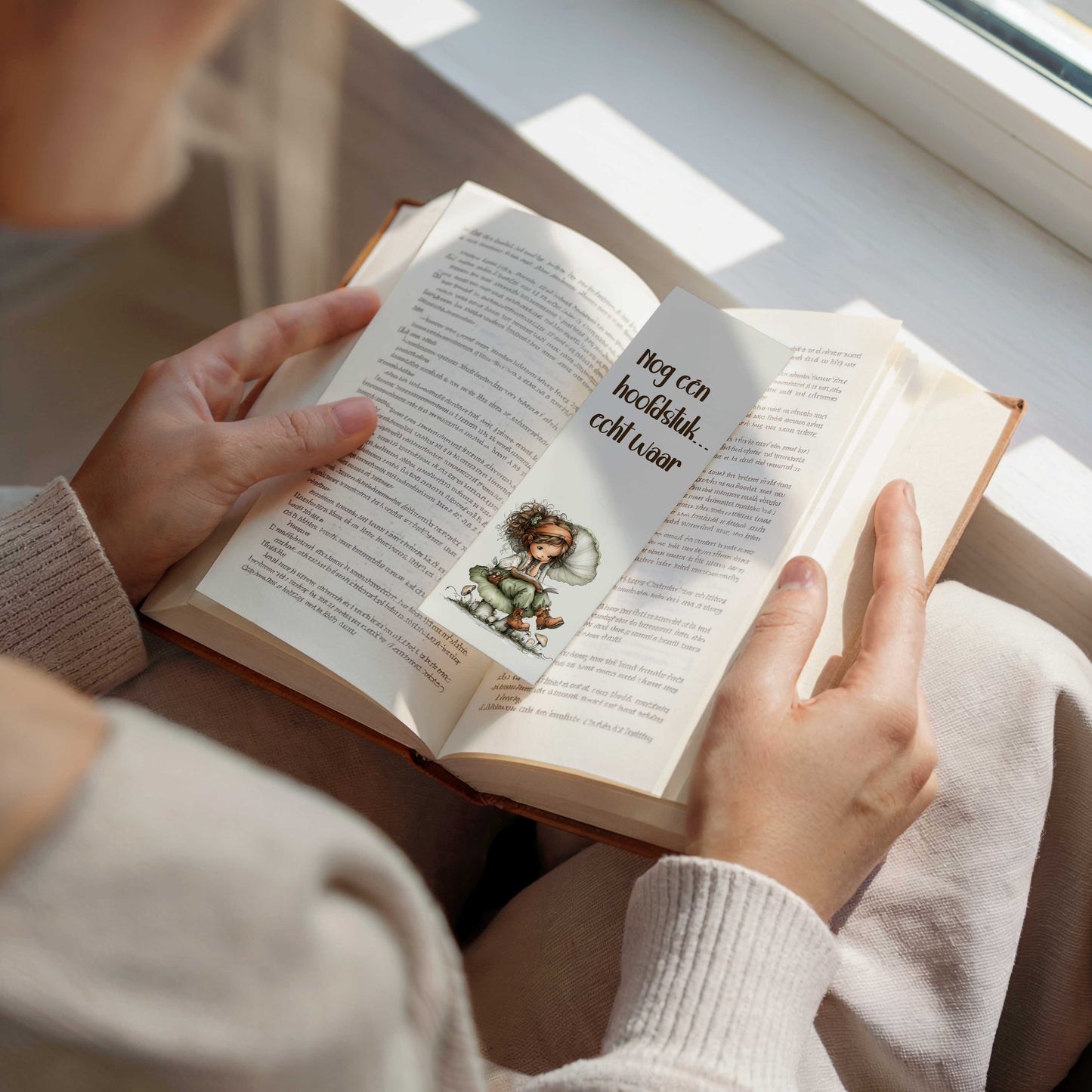 Boekenlegger met de tekst "Nog één hoofdstuk... echt waar!" en een fee die op een paddenstoel een boek leest, geplaatst tussen de pagina's van een open boek.