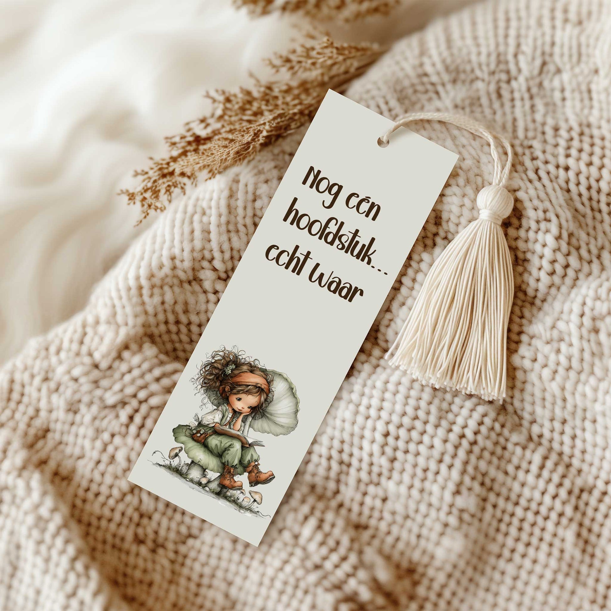 Boekenlegger met de tekst 'Nog één hoofdstuk... echt waar!' en een leesfee die op een paddenstoel zit te lezen. De boekenlegger met crèmekleurig kwastje ligt op een grof gebreide beige plaid met decoratieve droogbloemen.
