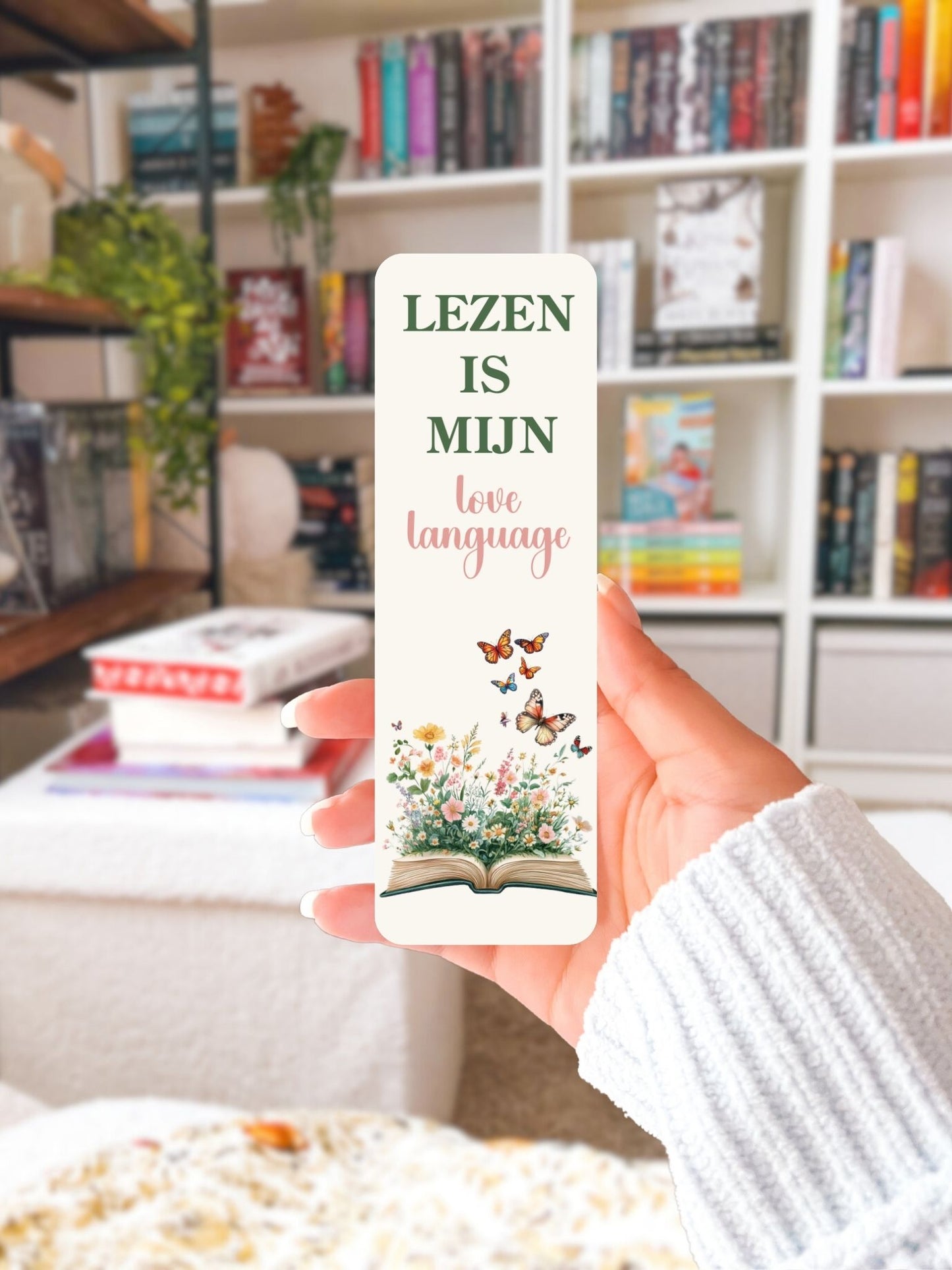 Hand houdt boekenlegger vast met bloemen en vlinders en de tekst Lezen is mijn love language, boekenkast op de achtergrond