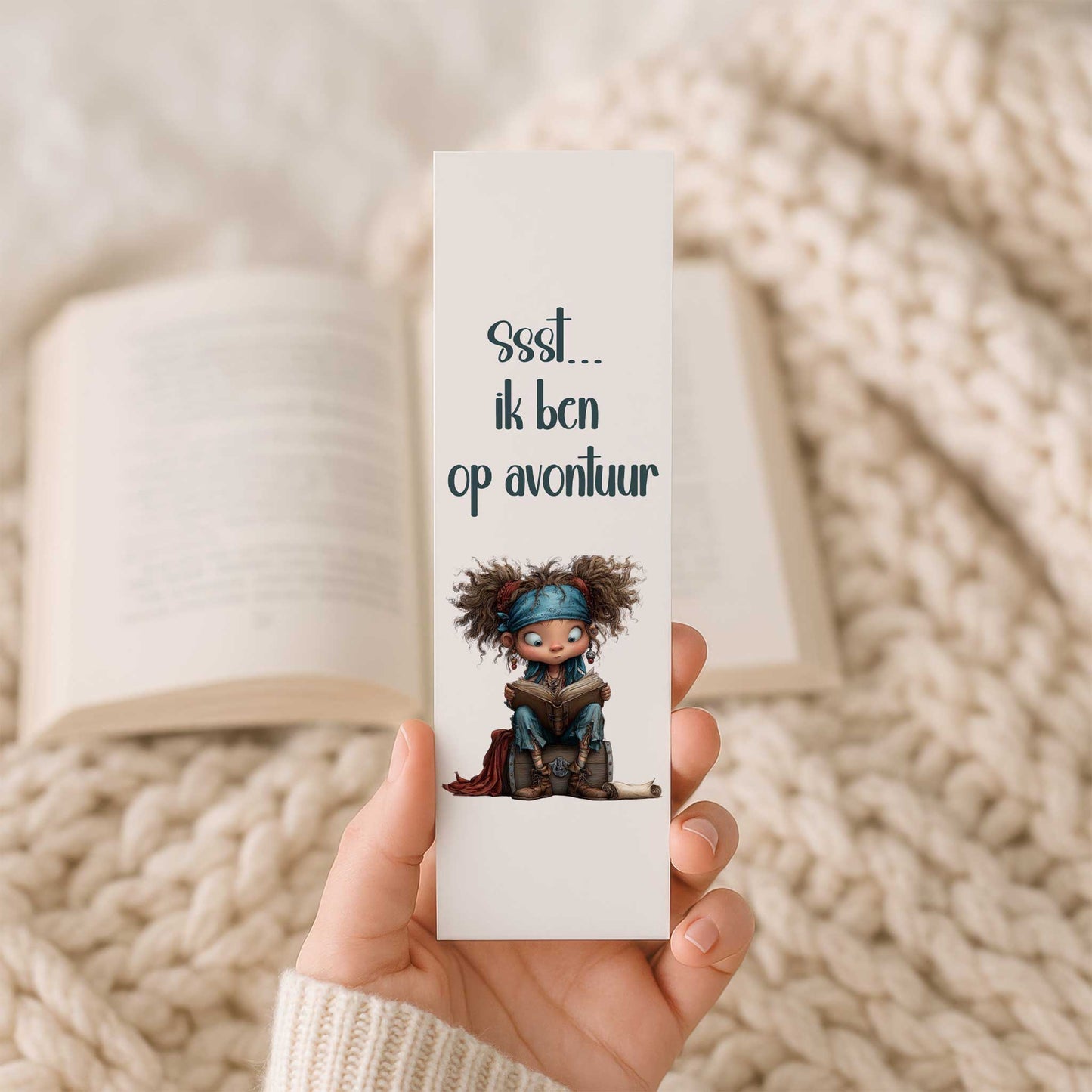Hand houdt een gelamineerde boekenlegger vast met een schattig lezend piratenkind op een schatkist en de tekst 'Ssst... ik ben op avontuur'. Op de achtergrond ligt een open boek.