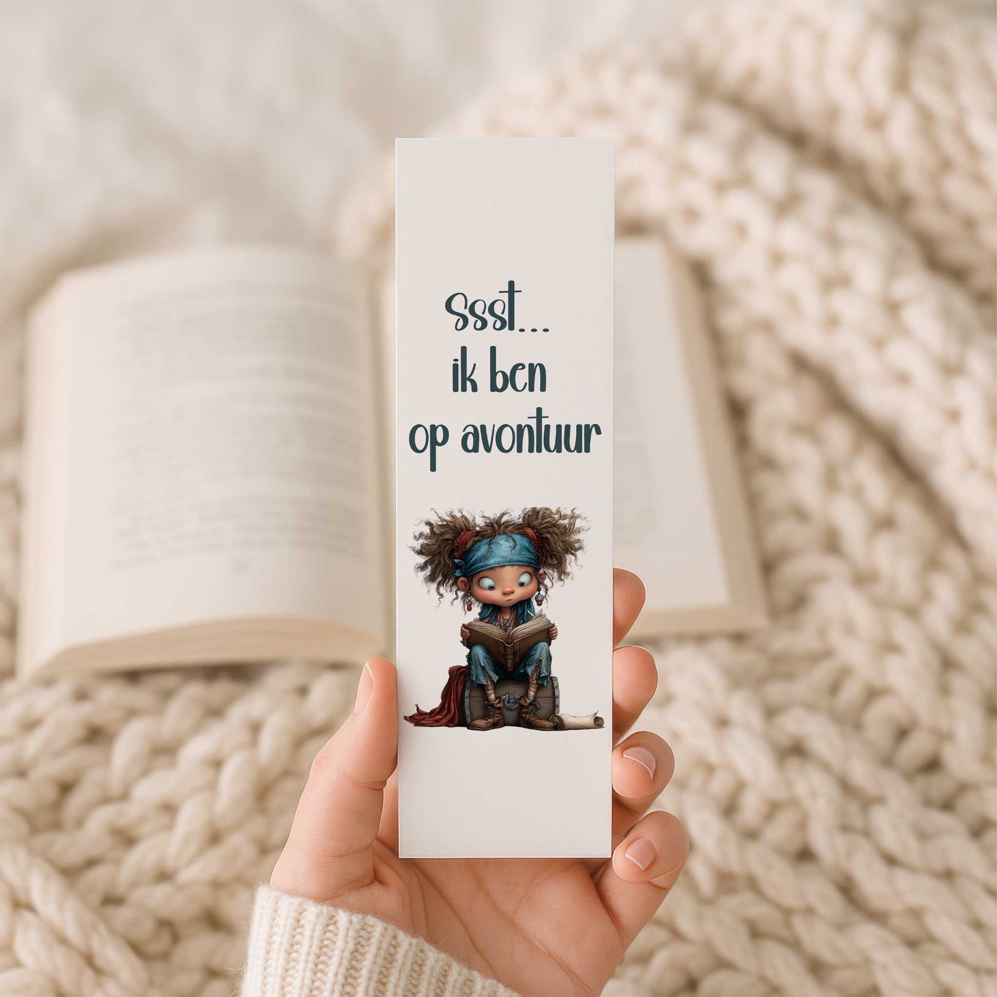Hand houdt een gelamineerde boekenlegger vast met een schattig lezend piratenkind op een schatkist en de tekst 'Ssst... ik ben op avontuur'. Op de achtergrond ligt een open boek.