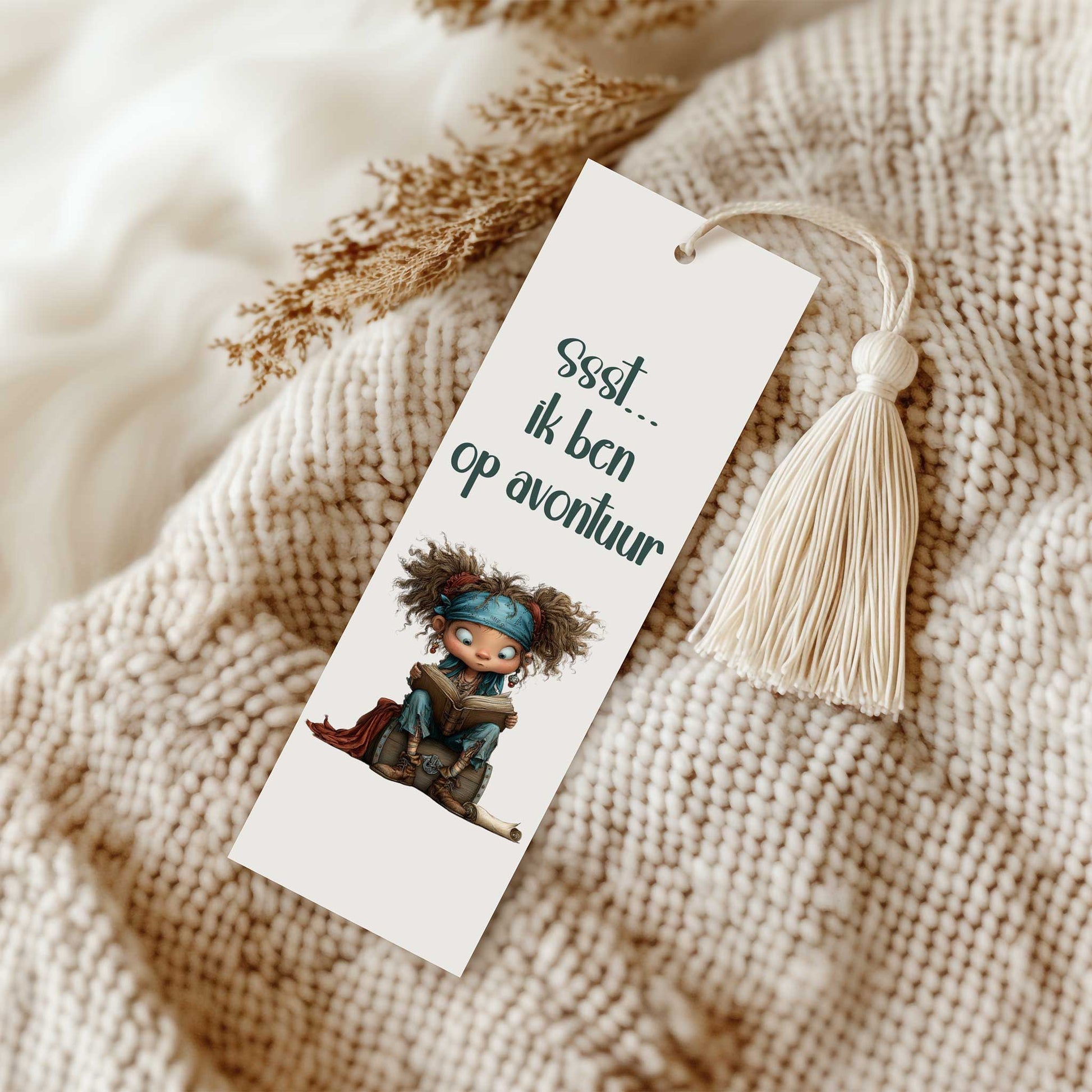 Gelamineerde boekenlegger met een crèmekleurig kwastje op een gebreide beige deken. Op de boekenlegger staat een lezend piratenkind en de tekst 'Ssst... ik ben op avontuur'.