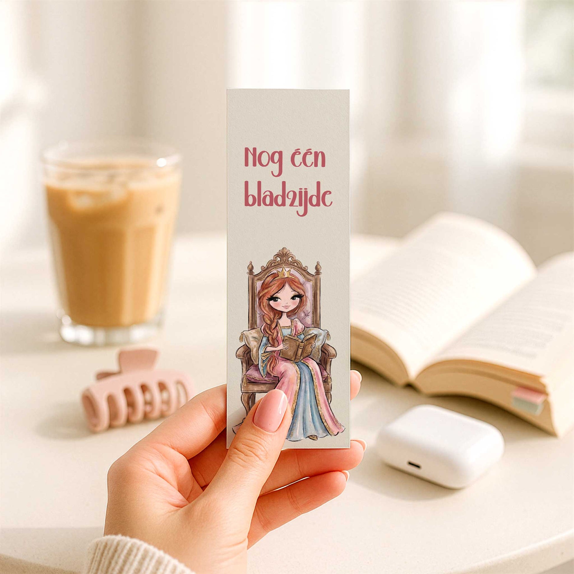 Hand houdt een boekenlegger vast met de tekst 'Nog één bladzijde' en een illustratie van een prinses die lezend op een sierlijke troon zit. Op de achtergrond staan een open boek, een glas ijskoffie en een witte oplaadcase.