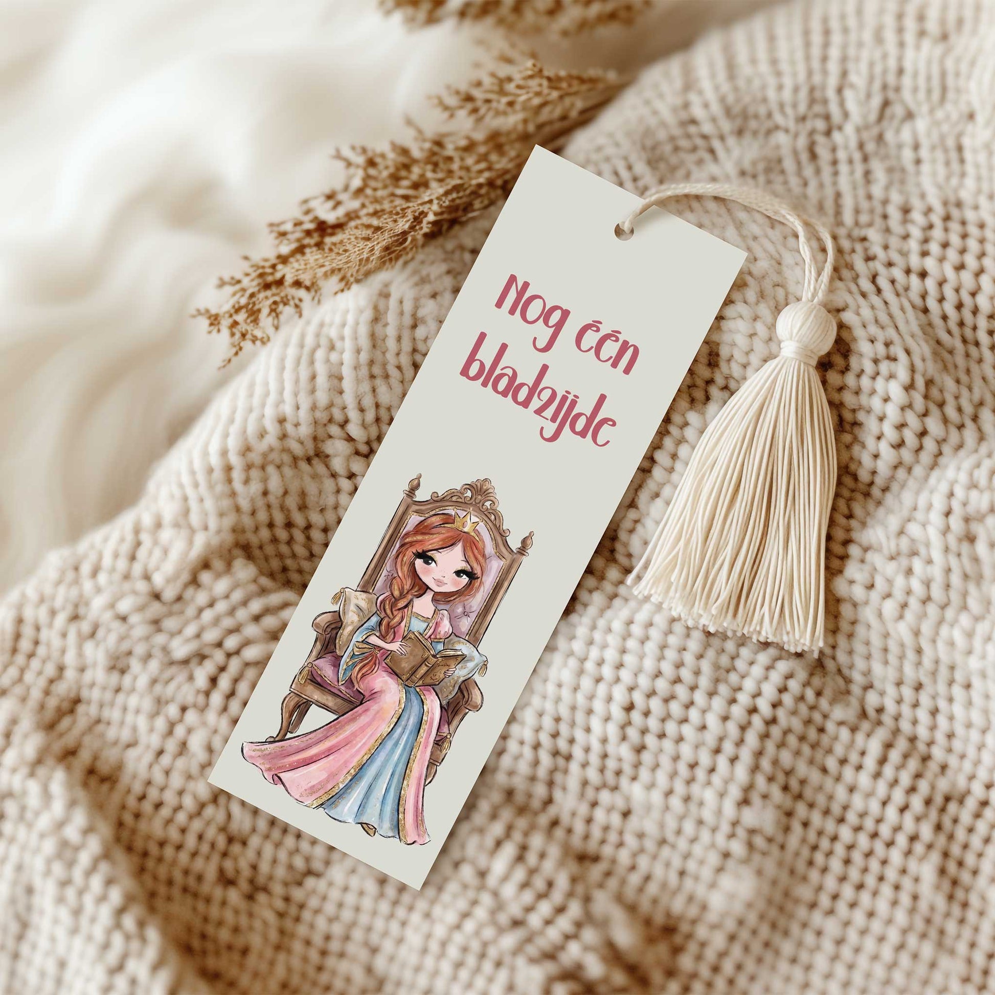 Boekenlegger met de tekst 'Nog één bladzijde' en een lezende prinses op een sierlijke troon. De boekenlegger ligt op een beige gebreide deken en heeft een crèmekleurig kwastje.