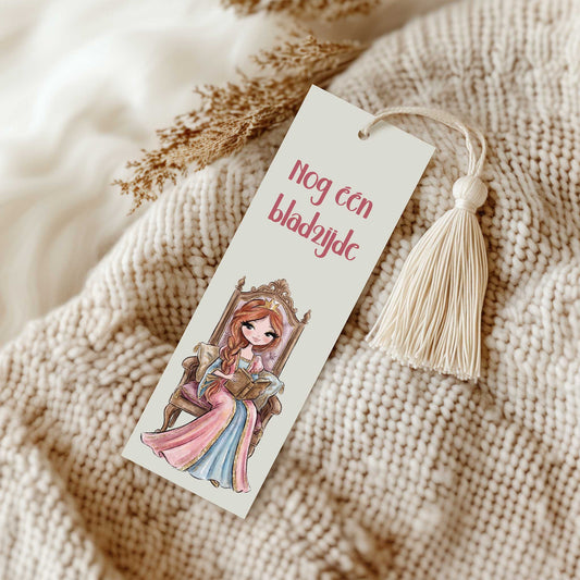 Boekenlegger met de tekst 'Nog één bladzijde' en een lezende prinses op een sierlijke troon. De boekenlegger ligt op een beige gebreide deken en heeft een crèmekleurig kwastje.