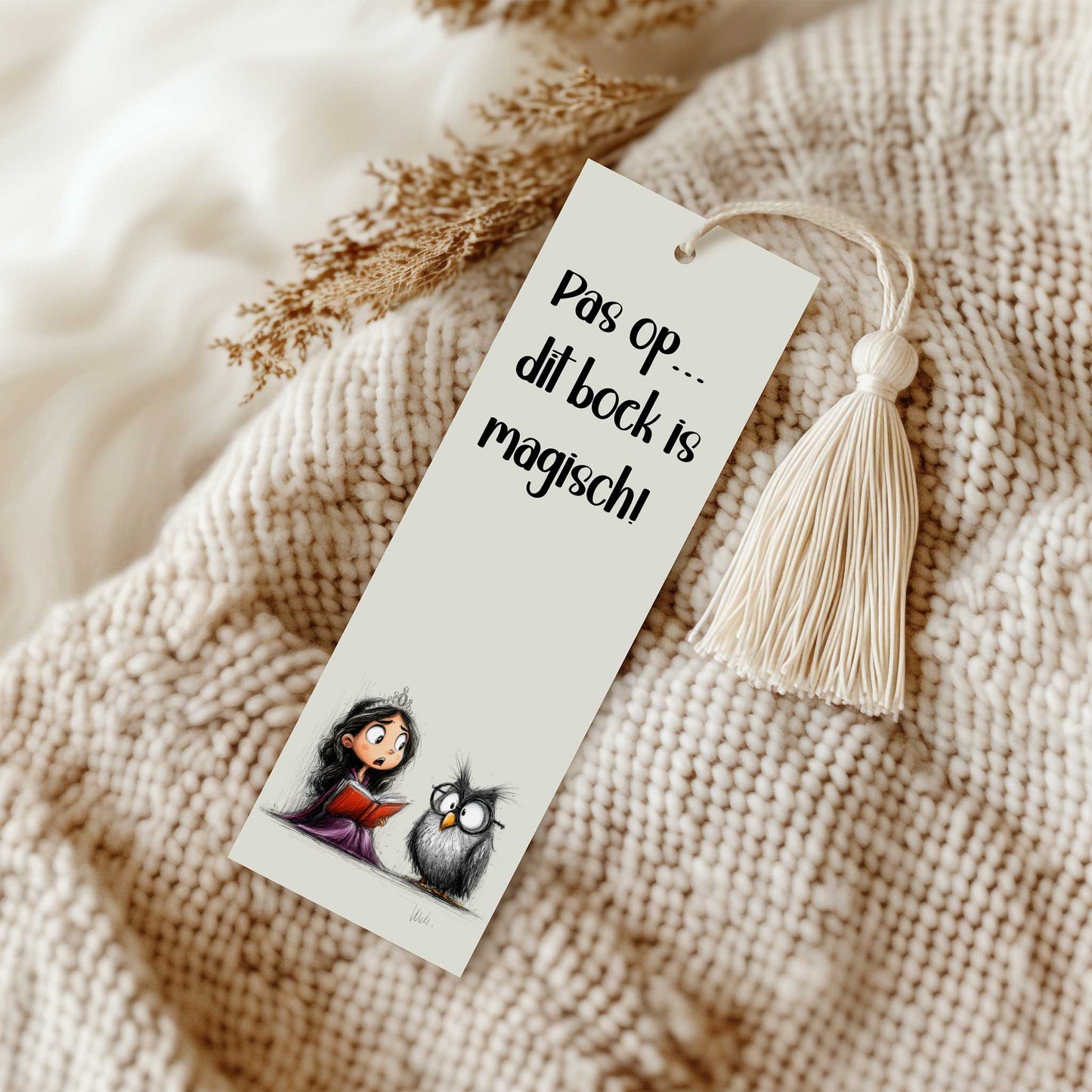 Boekenlegger met de tekst 'Pas op... dit boek is magisch!' en een illustratie van een prinses met een rood boek naast een uil. De boekenlegger ligt op een beige gebreide deken naast een crèmekleurig kwastje.