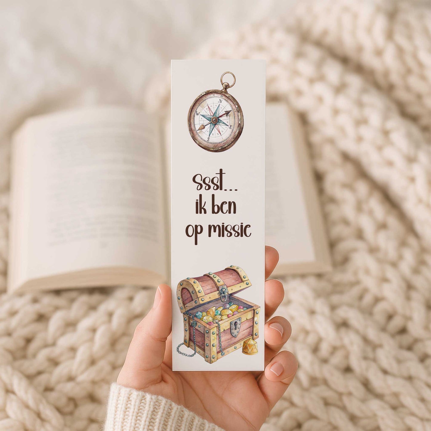 Productfoto van een hand die een boekenlegger vasthoudt met een kompas bovenaan, de tekst 'Ssst... ik ben op missie' en een open schatkist, met een open boek op de achtergrond.