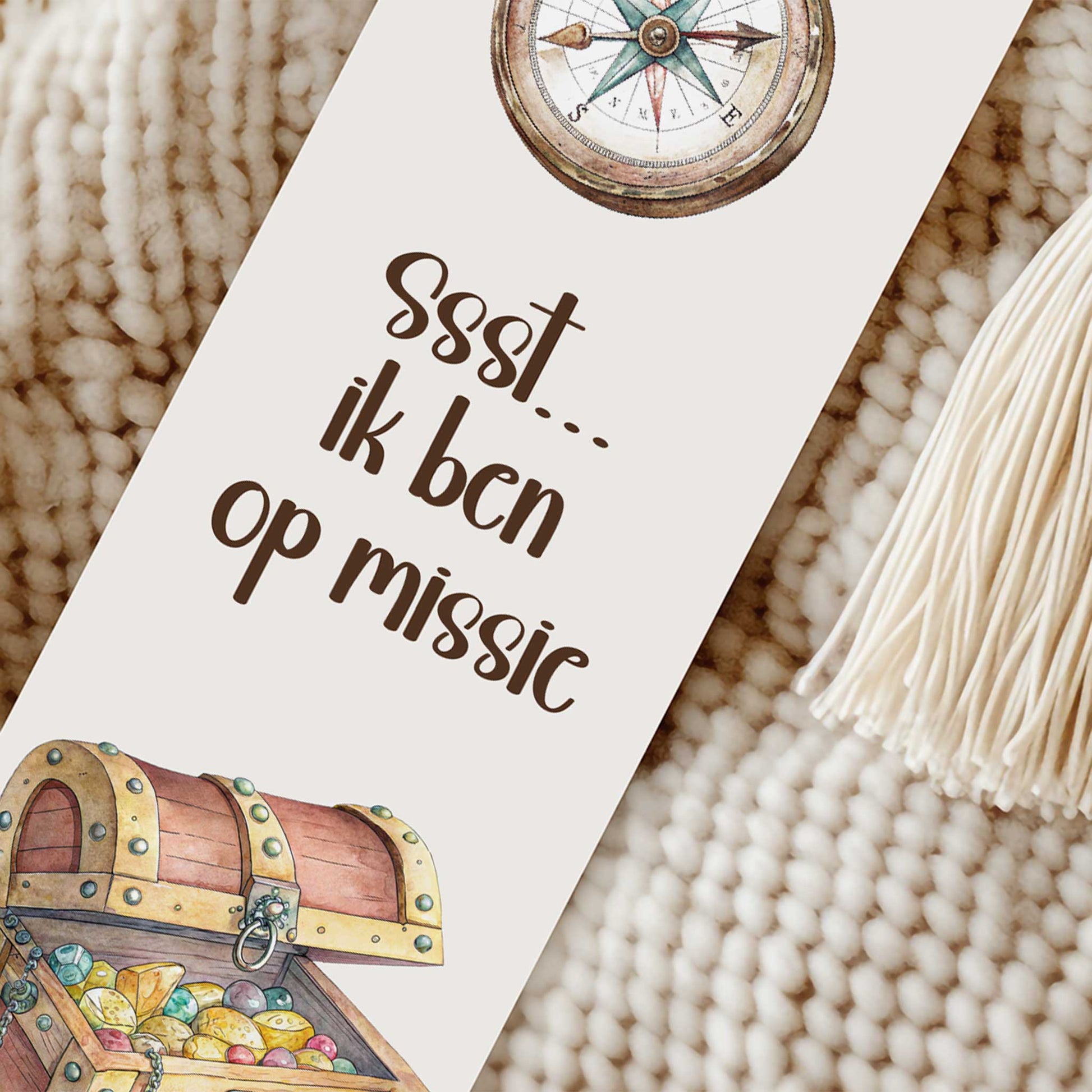 Close-up van een boekenlegger met een kompas bovenaan, de tekst 'Ssst... ik ben op missie', een open schatkist onderaan en een crèmekleurig kwastje op een gebreide beige ondergrond.