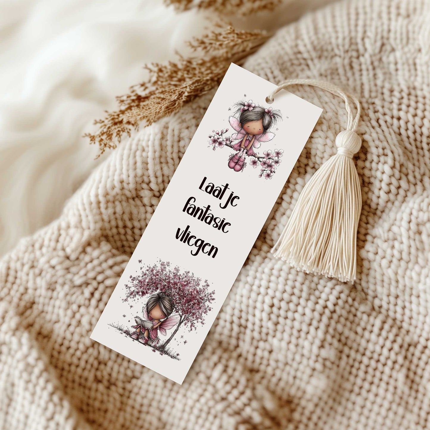 Boekenlegger met de tekst "Laat je fantasie vliegen" en twee roze fee-illustraties op een grof gebreide beige plaid, naast een crèmekleurig kwastje.