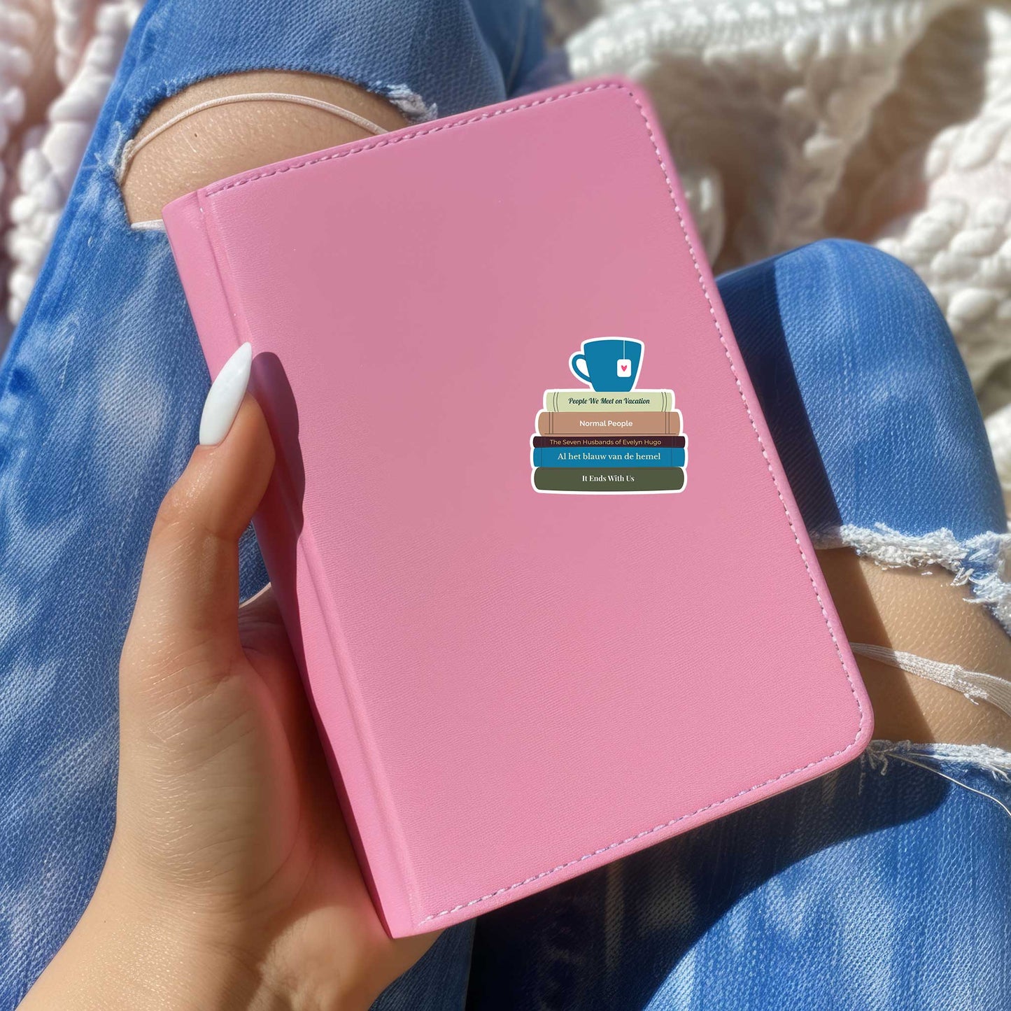Sticker met stapel boeken en theekopje geplakt op een roze boekje, vastgehouden door iemand in een blauwe ripped jeans. De sticker toont vijf populaire boektitels in diverse kleuren.