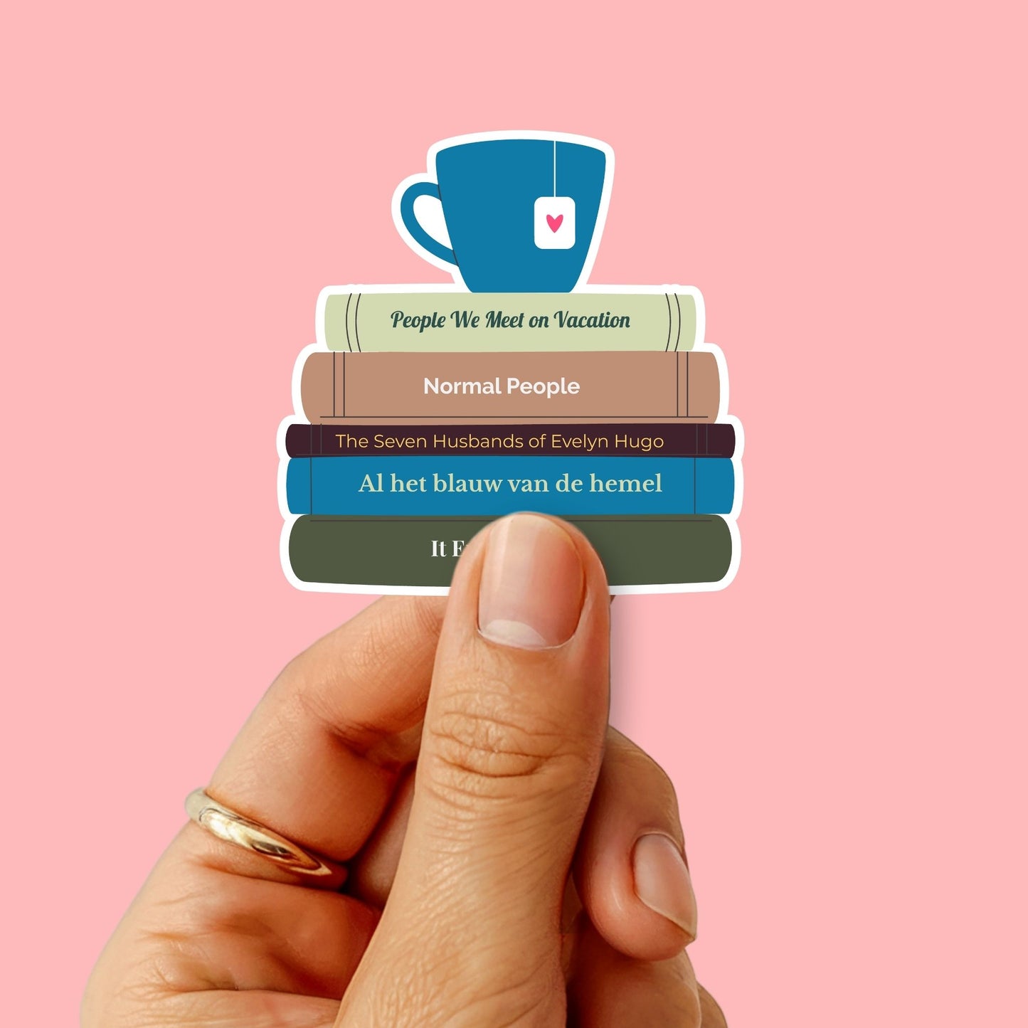 Hand houdt een kleurrijke boekenliefhebbers-sticker vast met een stapel boeken en een blauw theekopje erbovenop. De boeken zijn voorzien van titels zoals 'Normal People' en 'Al het blauw van de hemel'.