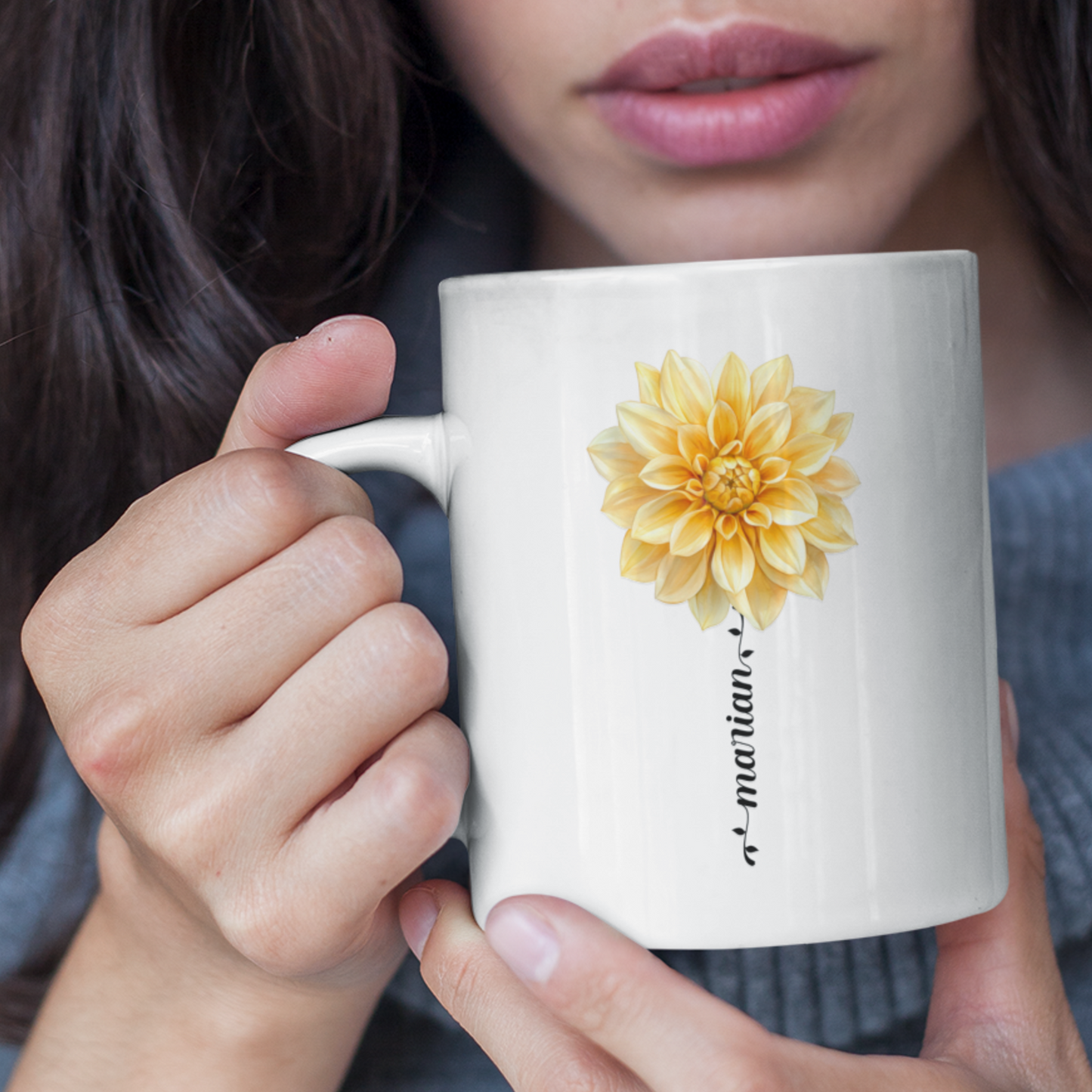 close up van een vrouw die een witte mok met gele Dahlia en personaliseerbare naam in haar hand houdt, naam is in sierlijke letters in de vorm van een bloemstengel in de kleur zwart. Van vrouw is deel van haar gezicht te zien.