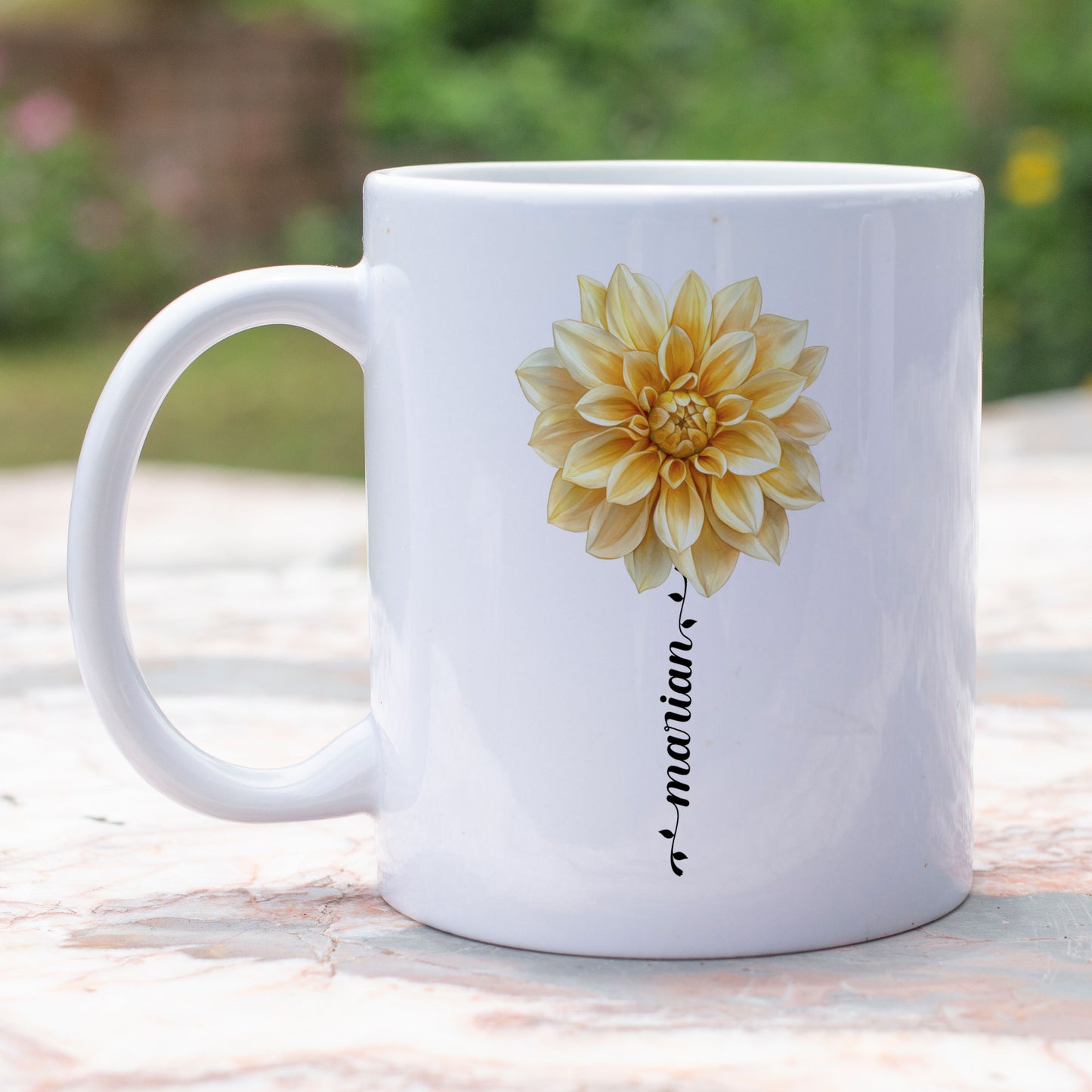 witte mok met gele Dahlia en personaliseerbare naam, naam is in sierlijke letters in de vorm van een bloemstengel in de kleur zwart. Mok staat buiten op een tuintafel