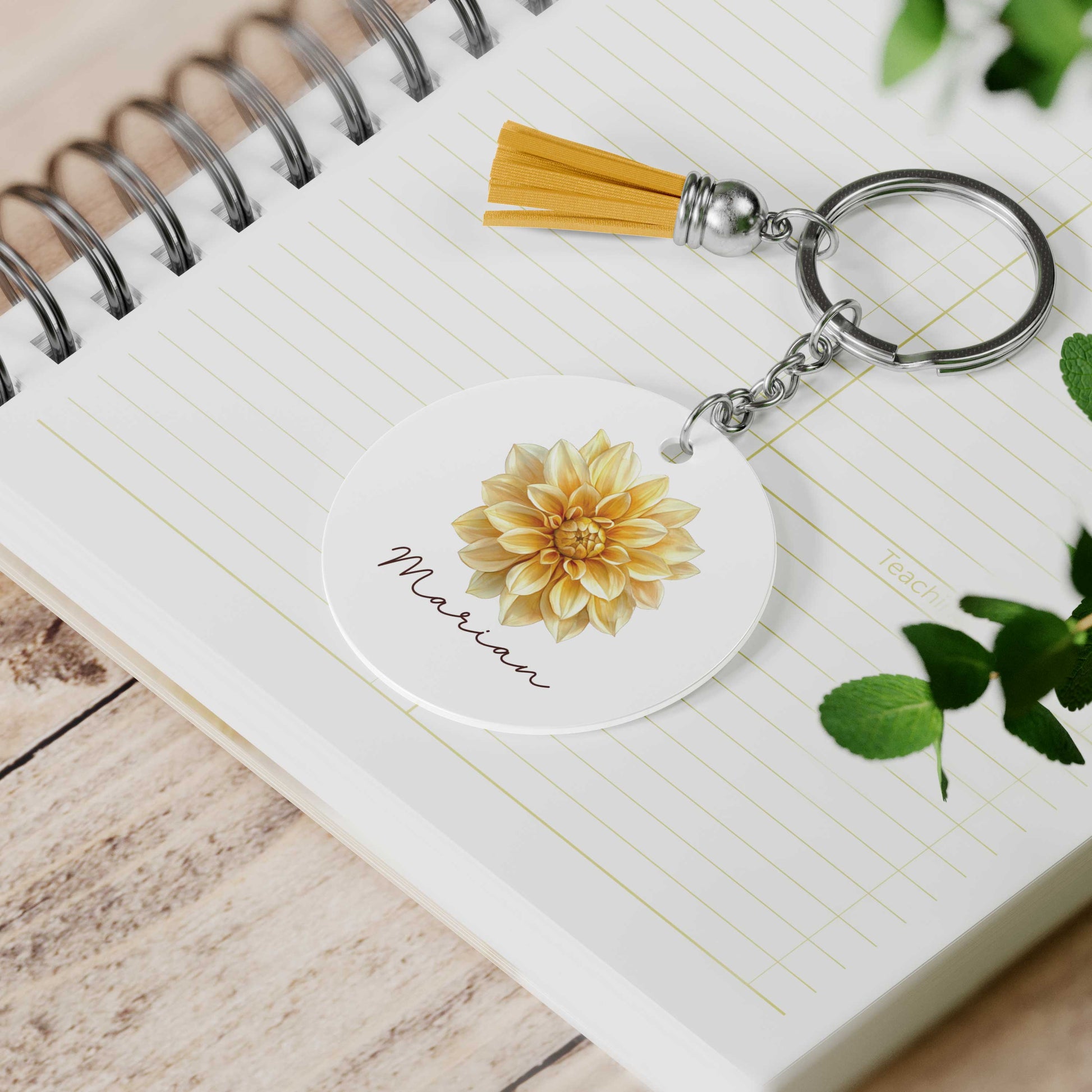Ronde sleutelhanger met een gele dahlia-illustratie en een gepersonaliseerde naam op een open notitieboek. met een geel kwastje