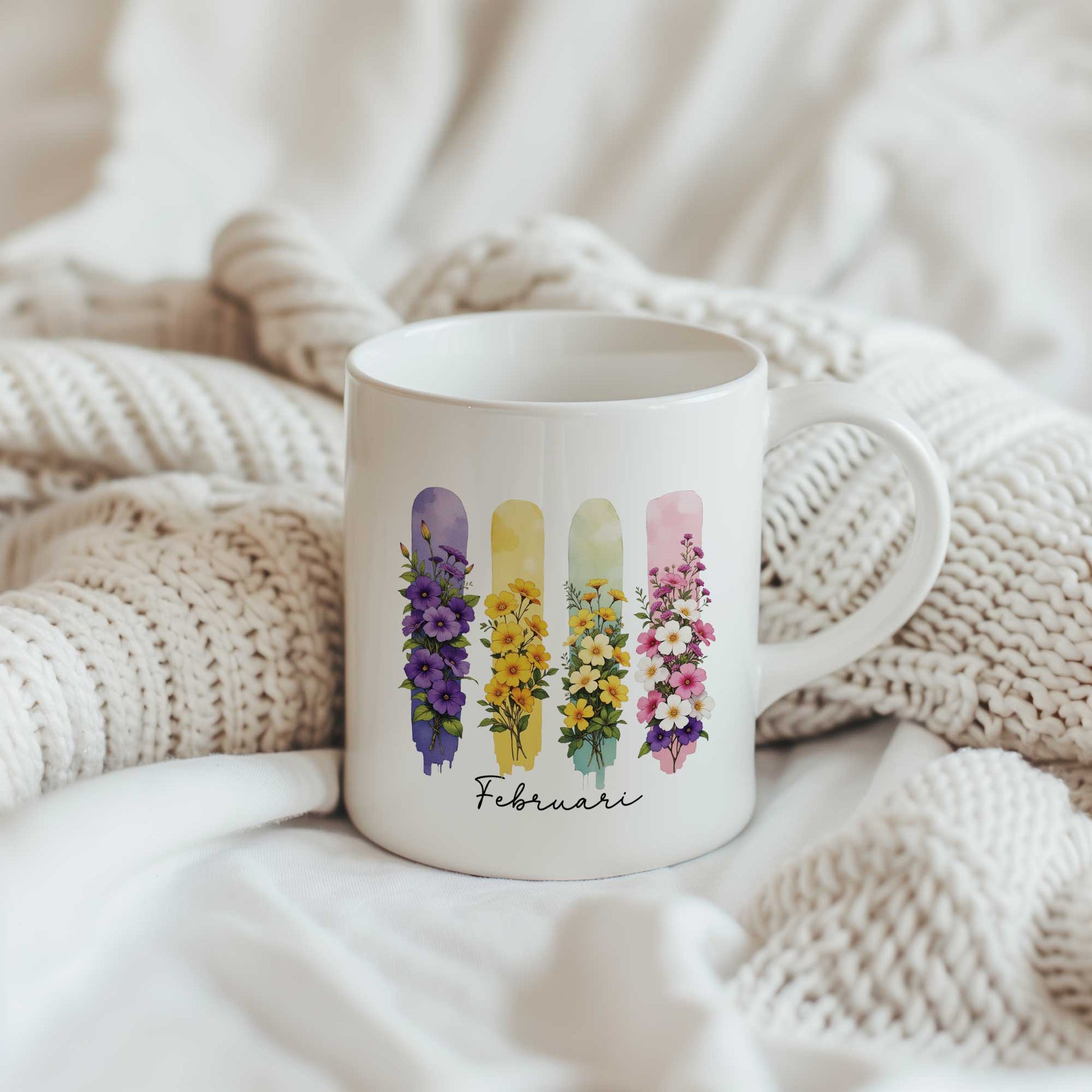 Witte mok met de geboortebloemen van februari in brush effect op een beige gebreide plaid, met het bloemenontwerp en de tekst 'Februari' zichtbaar.