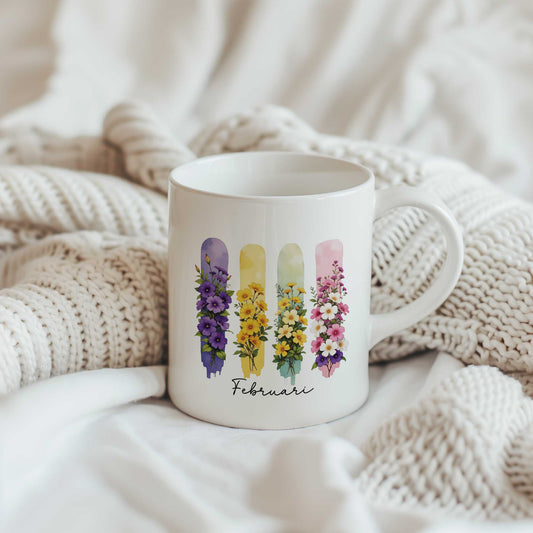 Witte mok met de geboortebloemen van februari in brush effect op een beige gebreide plaid, met het bloemenontwerp en de tekst 'Februari' zichtbaar.