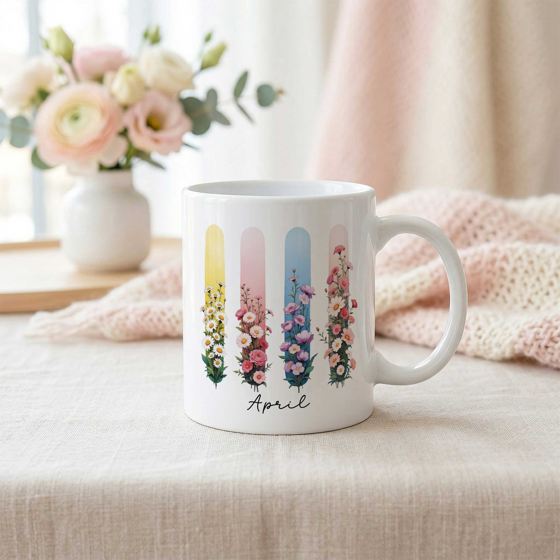 Witte mok met de tekst 'April' en een bloemenontwerp in brush effect op een licht linnen tafelkleed. Op de achtergrond staat een witte vaas met een boeket in zachte roze- en crèmetinten en ligt een zachtroze gebreide plaid.