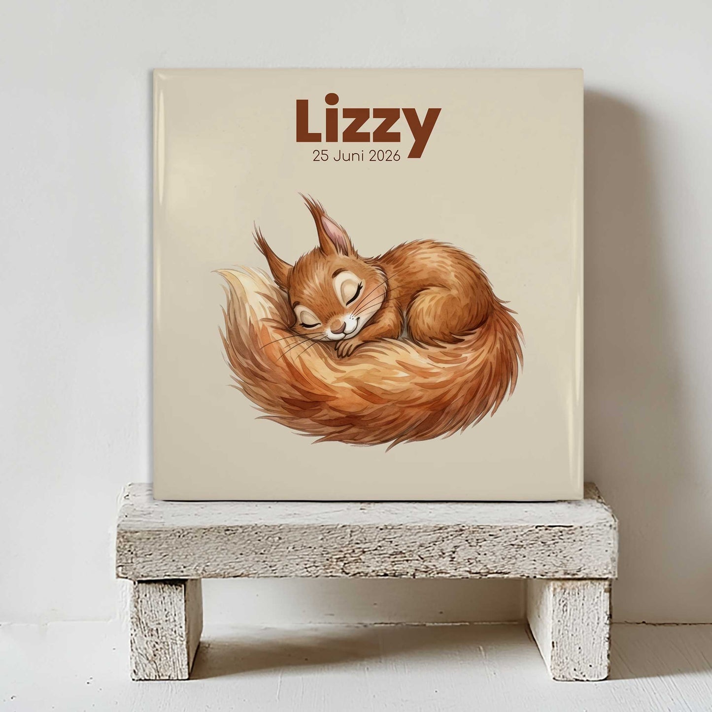 Keramisch babytegeltje met illustratie van slapend eekhoorntje, gepersonaliseerd met naam en geboortedatum, geplaatst op wit houten krukje tegen lichte muur.
