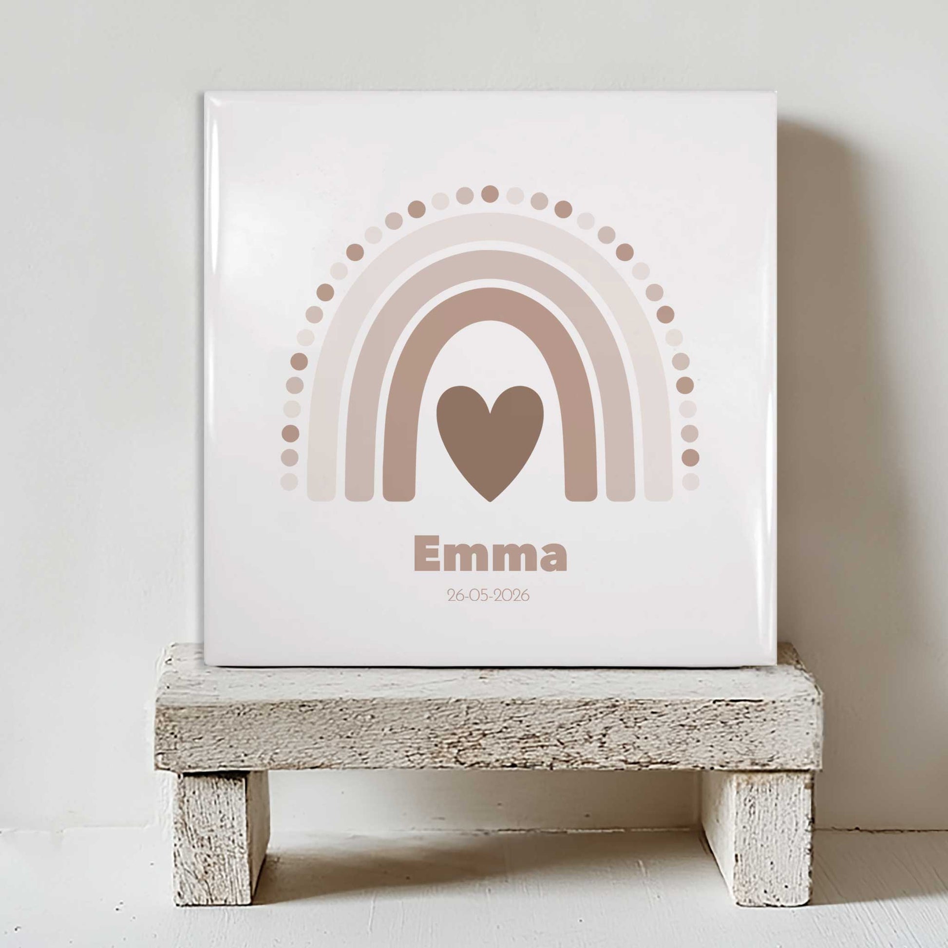 Vierkant keramisch geboortetegeltje met zachte regenboog in beige tinten en een hartje in het midden, gepersonaliseerd met de naam Emma en een datum, geplaatst op een klein houten krukje tegen een lichte muur