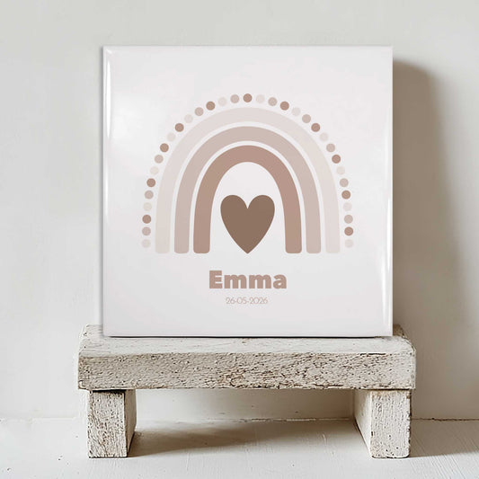 Vierkant keramisch geboortetegeltje met zachte regenboog in beige tinten en een hartje in het midden, gepersonaliseerd met de naam Emma en een datum, geplaatst op een klein houten krukje tegen een lichte muur