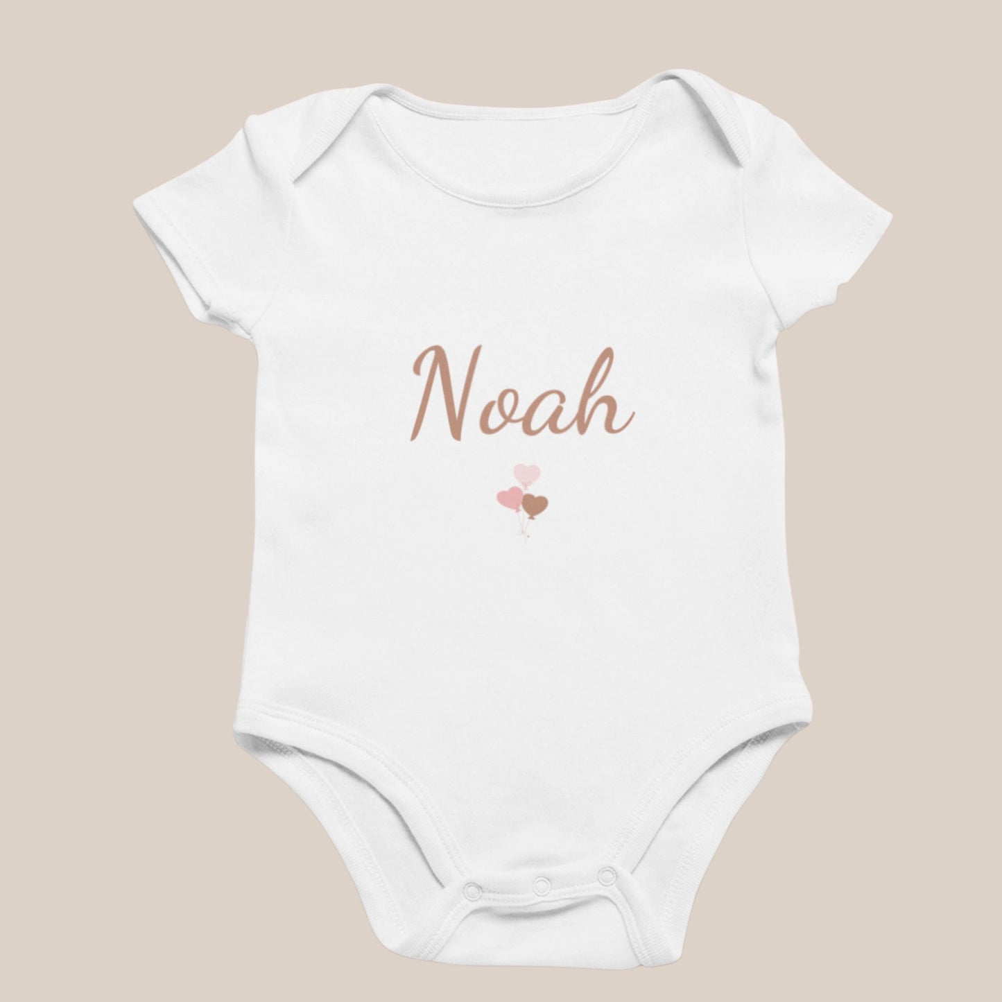 Wit baby rompertje met de naam Noah in zachte bruine letters en een klein blush hartjes ballonnen ontwerp eronder, weergegeven op een neutrale beige achtergrond.
