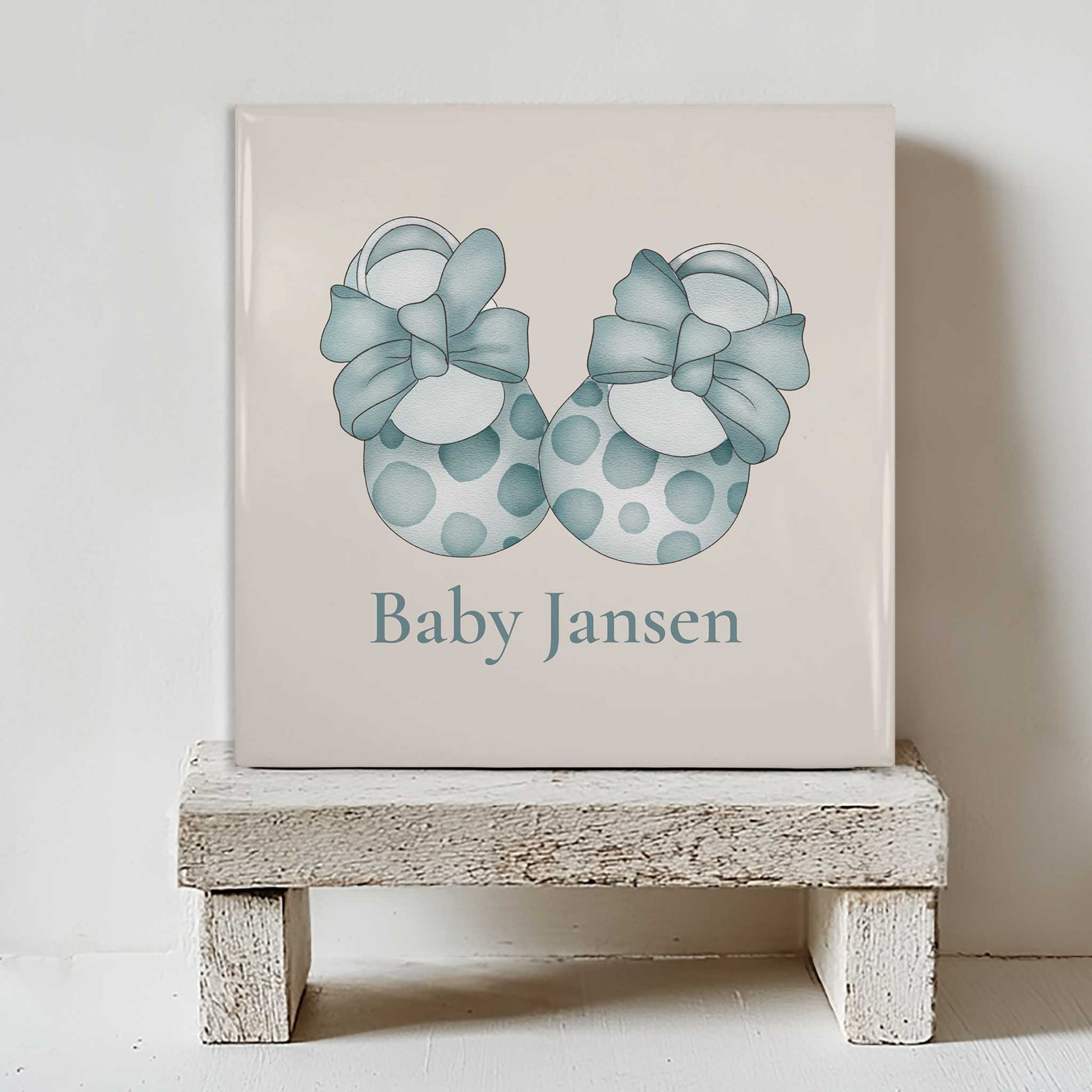 Gepersonaliseerd baby tegeltje met blauwe babyschoentjes en naam Baby Jansen geplaatst op een klein wit houten krukje tegen een lichte muur.
