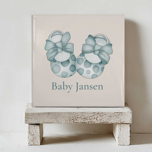 Gepersonaliseerd baby tegeltje met blauwe babyschoentjes en naam Baby Jansen geplaatst op een klein wit houten krukje tegen een lichte muur.