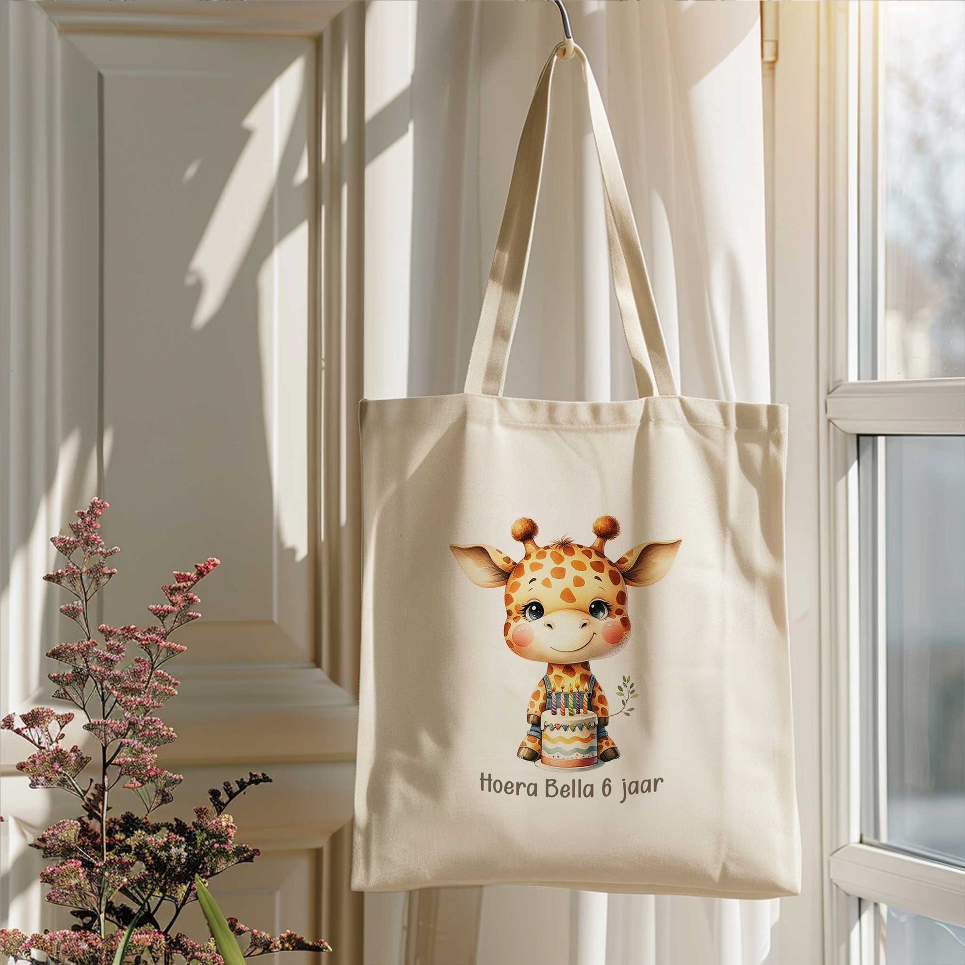 Gepersonaliseerde totebag met giraf en verjaardagstaart, bedrukt met de tekst 'Hoera Bella 6 jaar', hangend bij een raam in zacht daglicht.