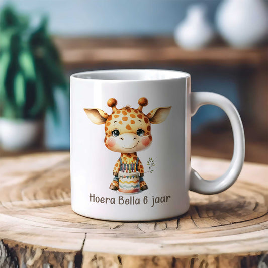 Witte gepersonaliseerde mok met giraf en verjaardagstaart op een houten tafel, bedrukt met de tekst 'Hoera Bella 6 jaar', met een zachte onscherpe achtergrond.
