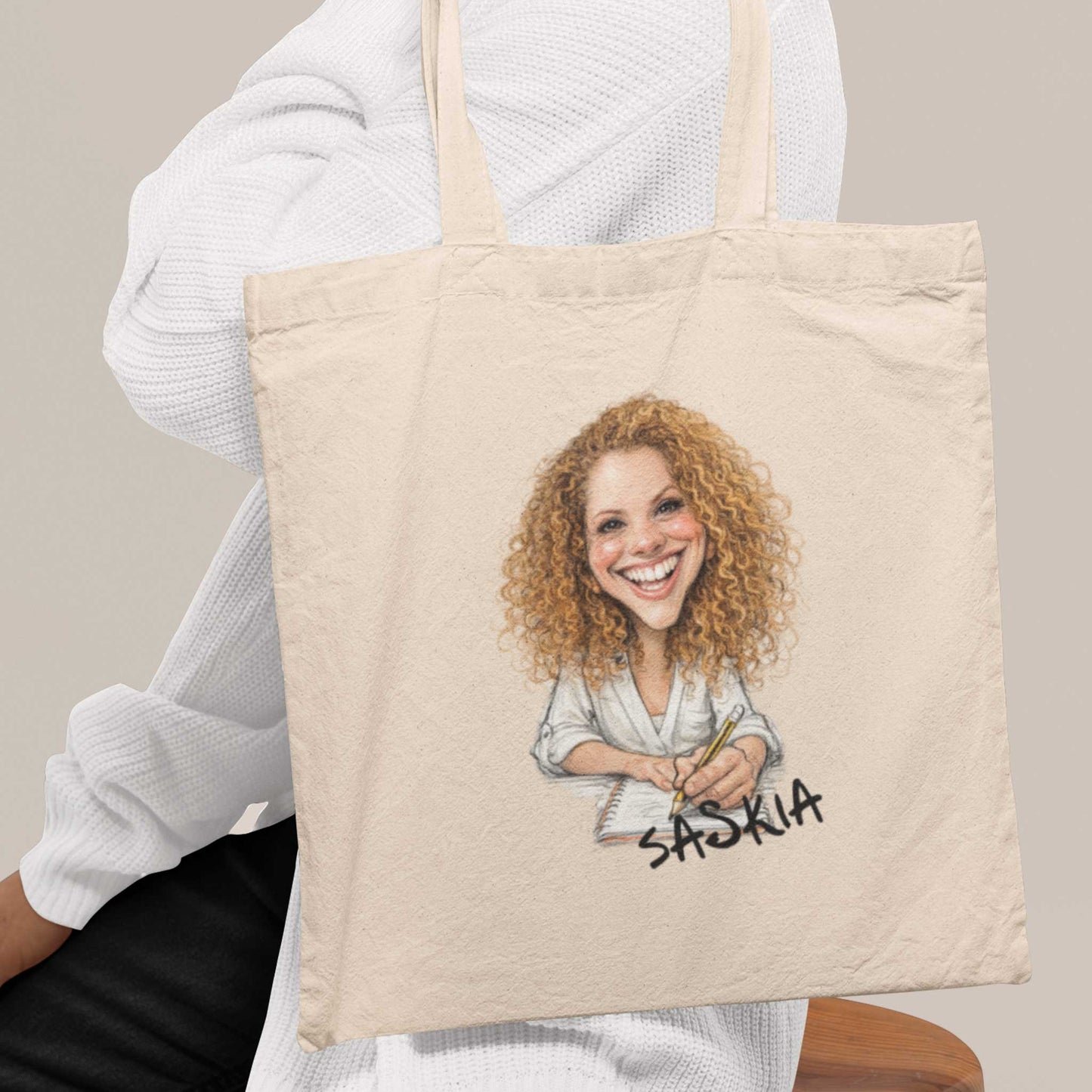 Vrouw in witte trui met beige katoenen totebag bedrukt met gepersonaliseerde karikatuur van een roodharige vrouw met krullen en de naam Saskia op de tas