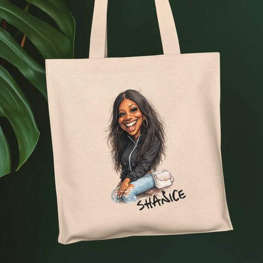 Beige katoenen totebag met gepersonaliseerde karikatuur illustratie van jonge vrouw met naam Shanice, gefotografeerd voor een donkere groene achtergrond met grote bladeren.