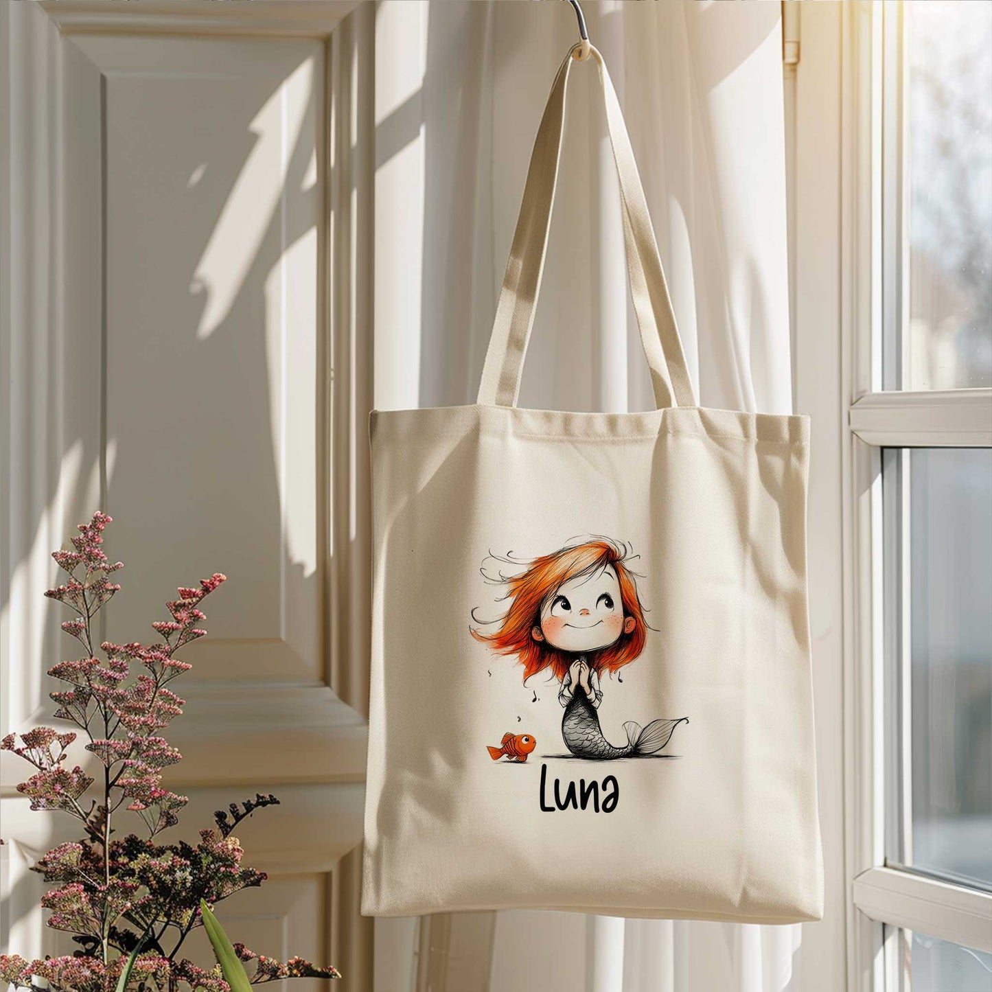 Gepersonaliseerde zeemeermin totebag met naam hangend bij een raam in een lichte kamer met bloemen op de voorgrond.