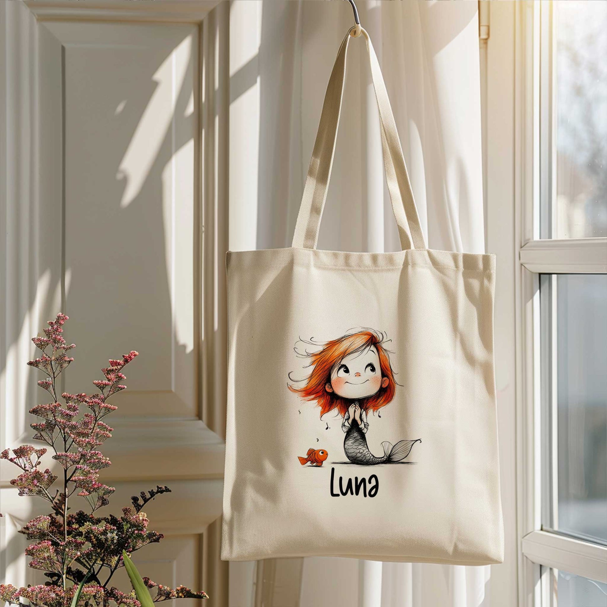 Gepersonaliseerde zeemeermin totebag met naam hangend bij een raam in een lichte kamer met bloemen op de voorgrond.