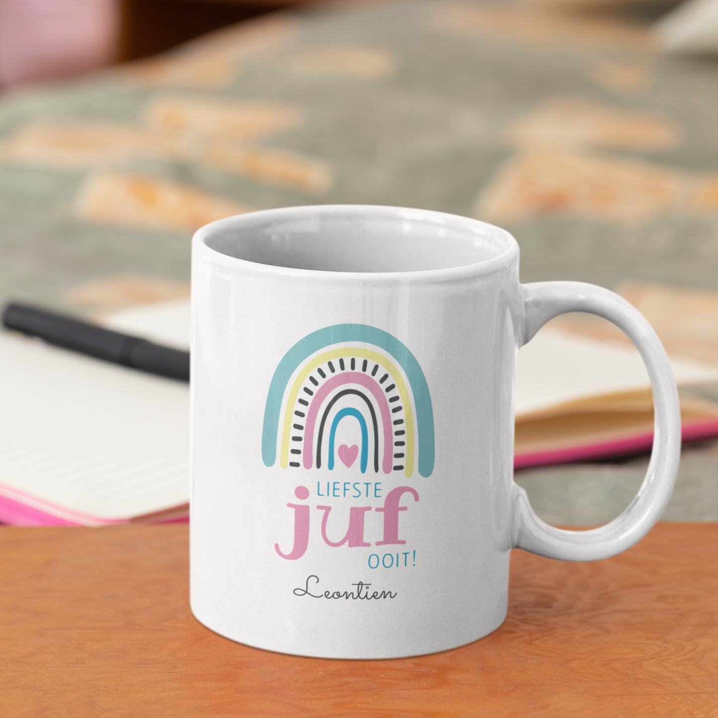 gepersonaliseerdewitte keramische mok voor juf met  boho regenboog en de tekst liefste juf ooit met naam  op een tafel naast een atlas