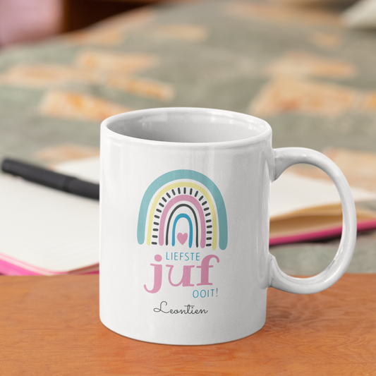 gepersonaliseerdewitte keramische mok voor juf met  boho regenboog en de tekst liefste juf ooit met naam  op een tafel naast een atlas