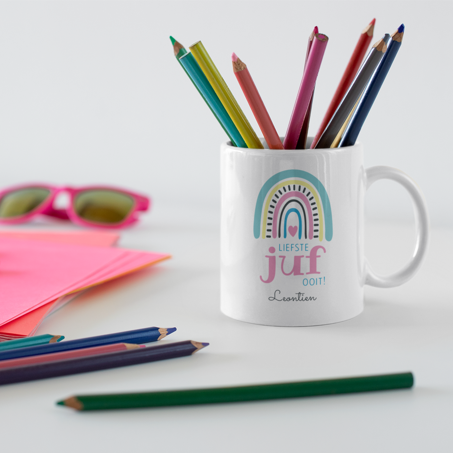 gepersonaliseerdewitte keramische mok voor juf met  boho regenboog en de tekst liefste juf ooit met naam op een tafel met kleurpotloden en roze vellen papier