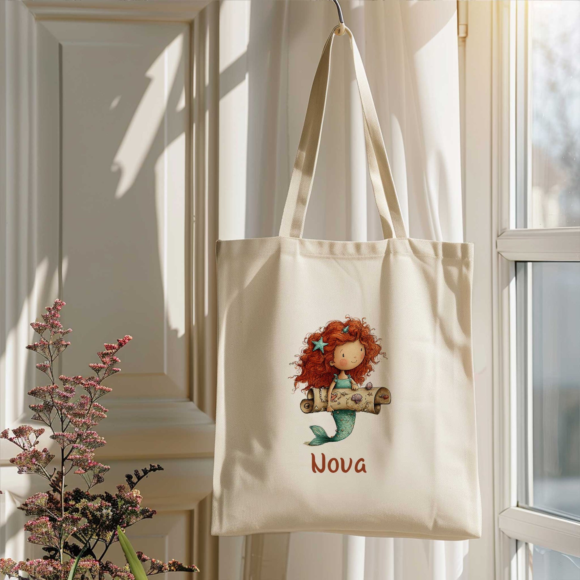 Zeemeermin totebag met naam Nova hangend bij een raam in zacht daglicht met bloemen op de voorgrond.