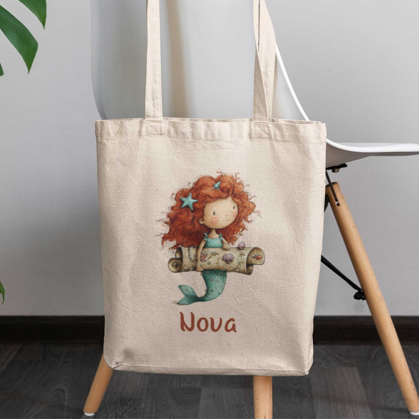 Zeemeermin totebag met naam Nova, hangend aan een stoel, met illustratie van een roodharige zeemeermin die een schatkaart vasthoudt.