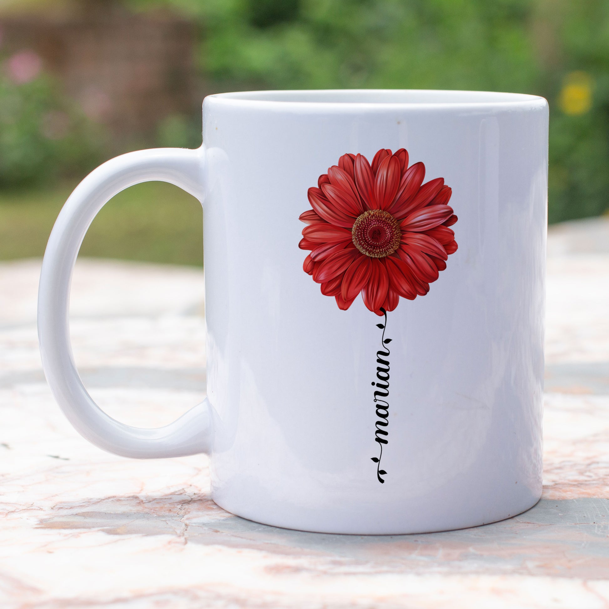 witte mok met rode gerbera en naam in sierlijk letterteken in de vorm van een  buiten op een tuintafel