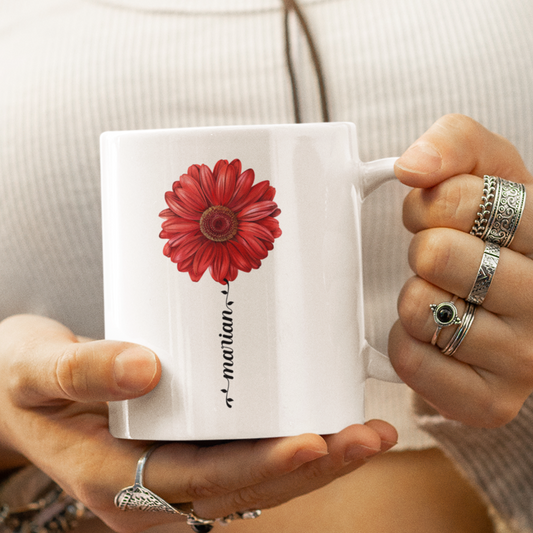 een vrouw me een witte mok in haar hand, een witte mok met rode gerbera en naam in sierlijk letterteken in de vorm van een bloemstengel
