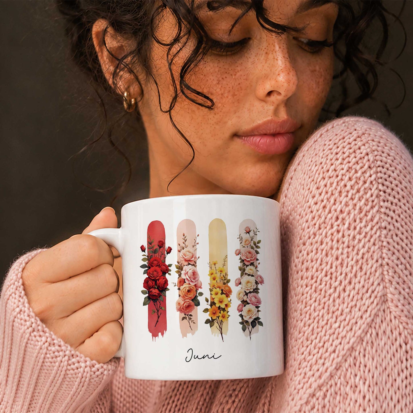 Vrouw in een roze gebreide trui houdt een witte mok vast met de geboortebloemen van juni.