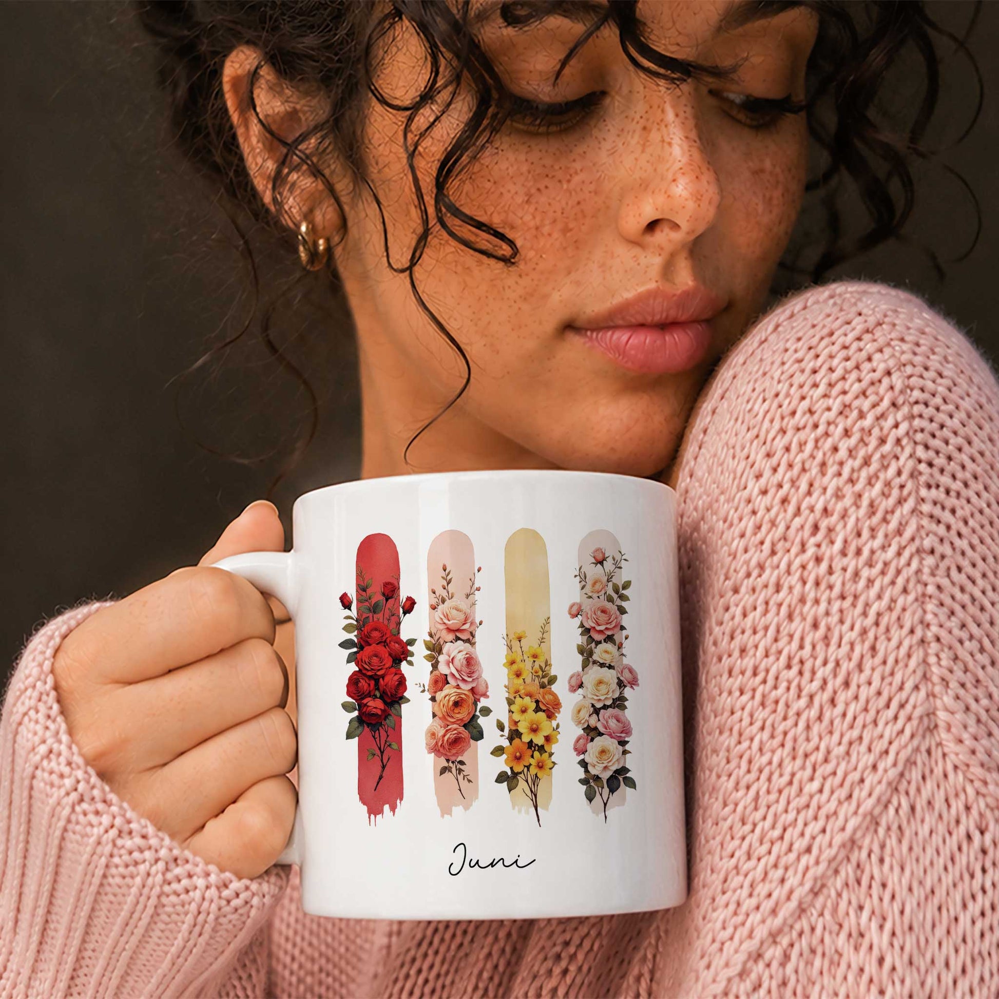 Vrouw in een roze gebreide trui houdt een witte mok vast met de geboortebloemen van juni.