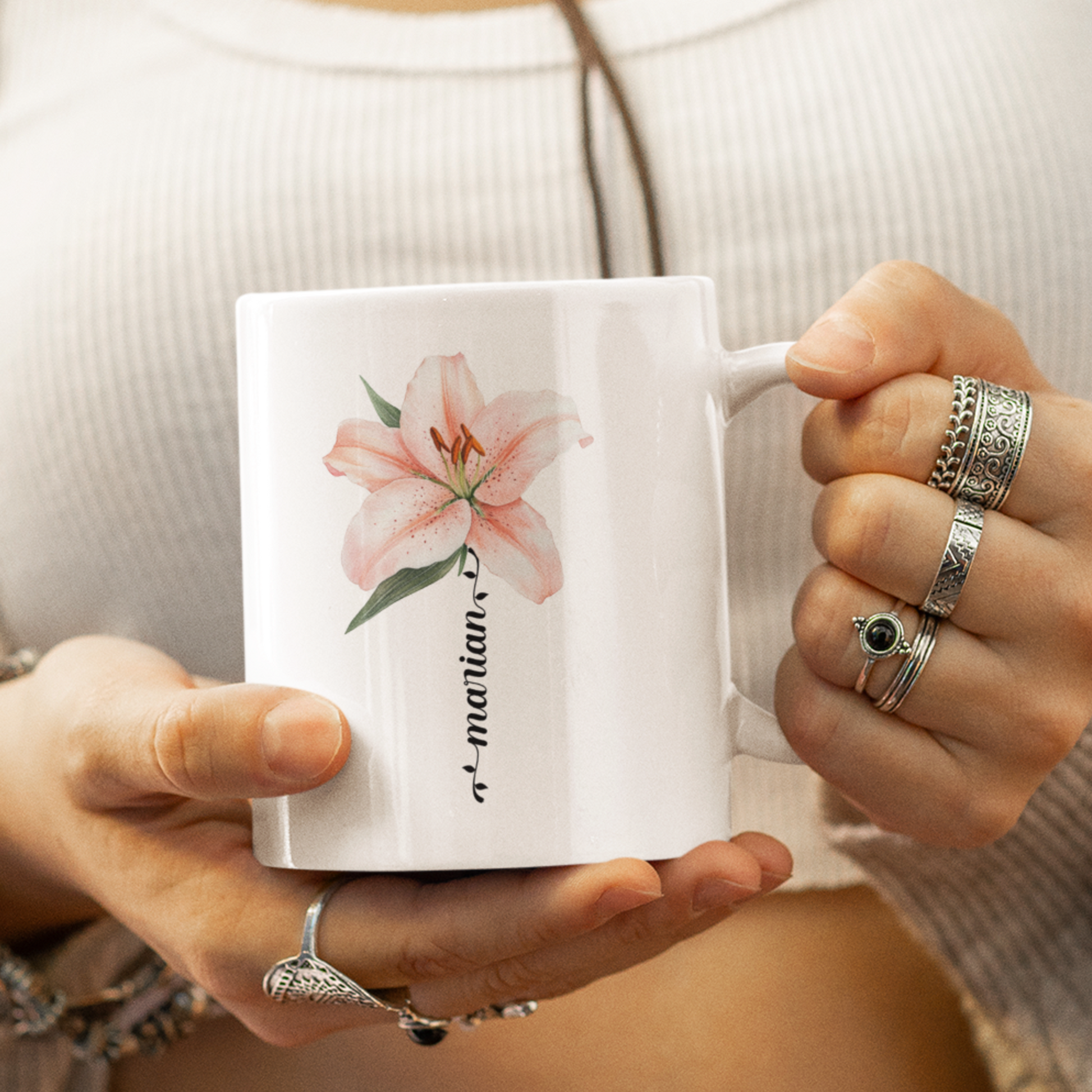 een vrouw met een mok in haar hand- met ringen om haar vingers, witte mok met licht roze lelie en personaliseerbare naam in sierlijke letters in de vorm van een bloemstengel in het zwart