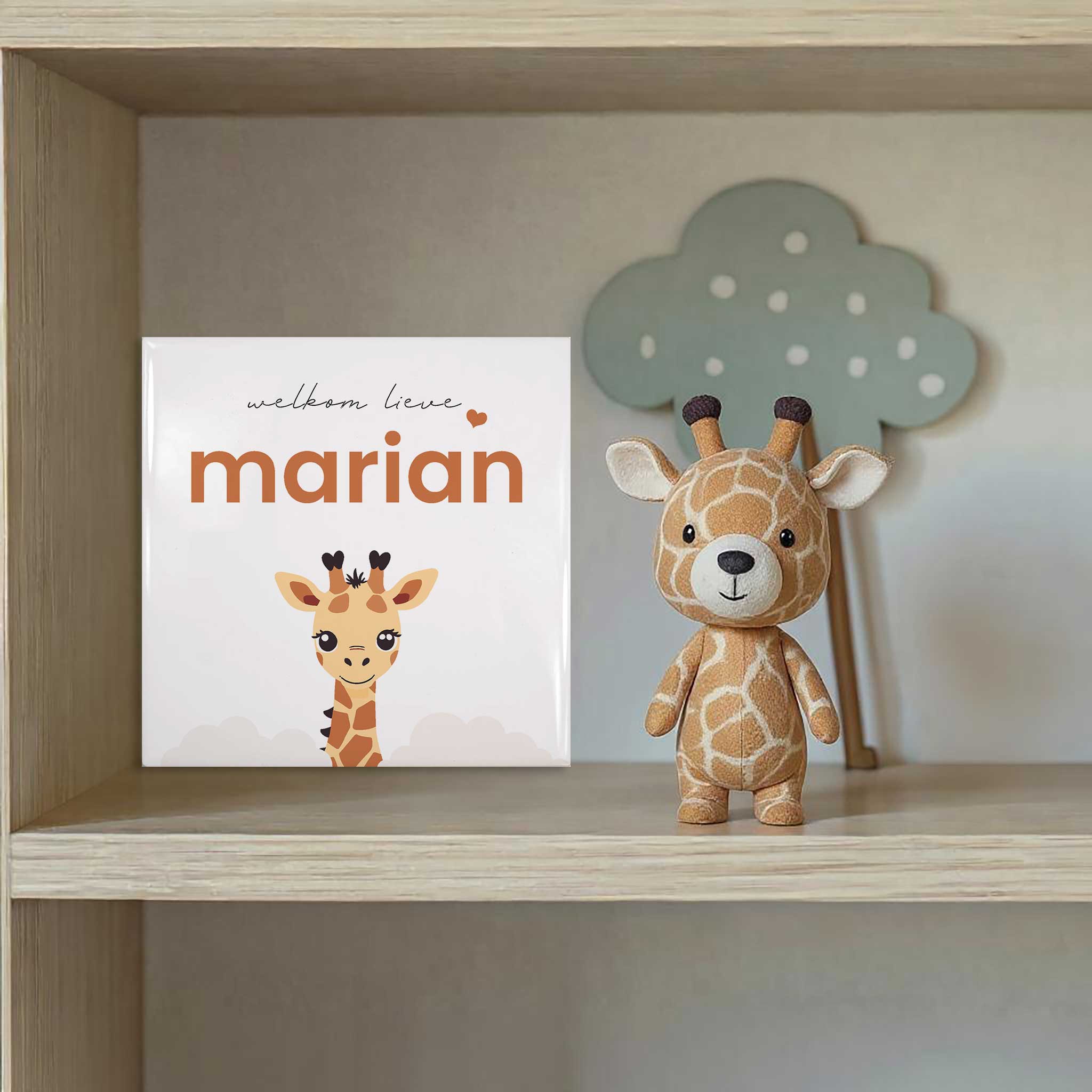 Lifestylefoto van een gepersonaliseerd geboortetegeltje met giraf en de naam Marian, op een plankje naast een giraffe-knuffel in een babykamer.
