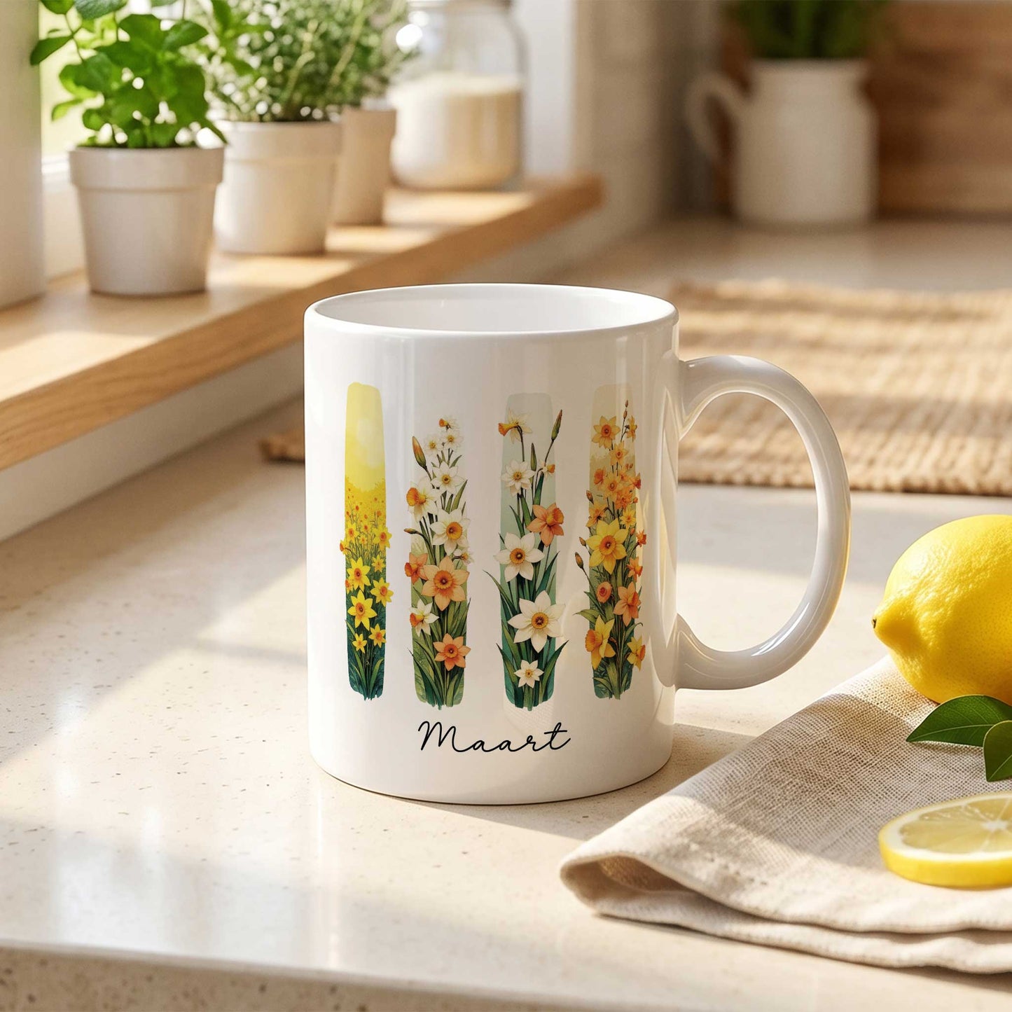 Witte mok met brush effect van gele en witte narcissen, de geboortebloem van maart, op een lichte keukenaanrecht met planten op de vensterbank en een citroen naast de mok.