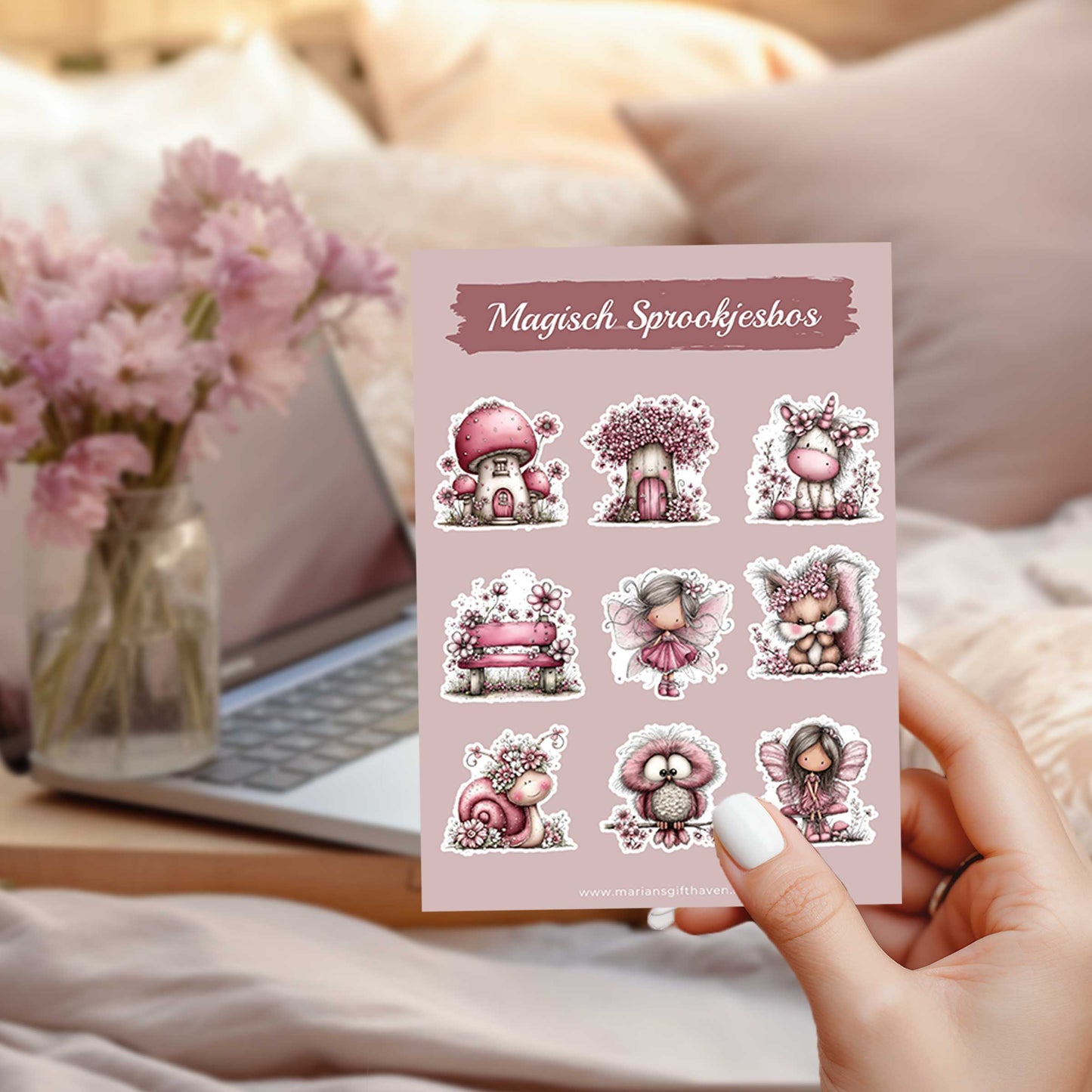 Hand houdt het stickervel Magisch Sprookjesbos vast boven een bed met op de achtergrond een laptop en een vaas met roze bloemen.