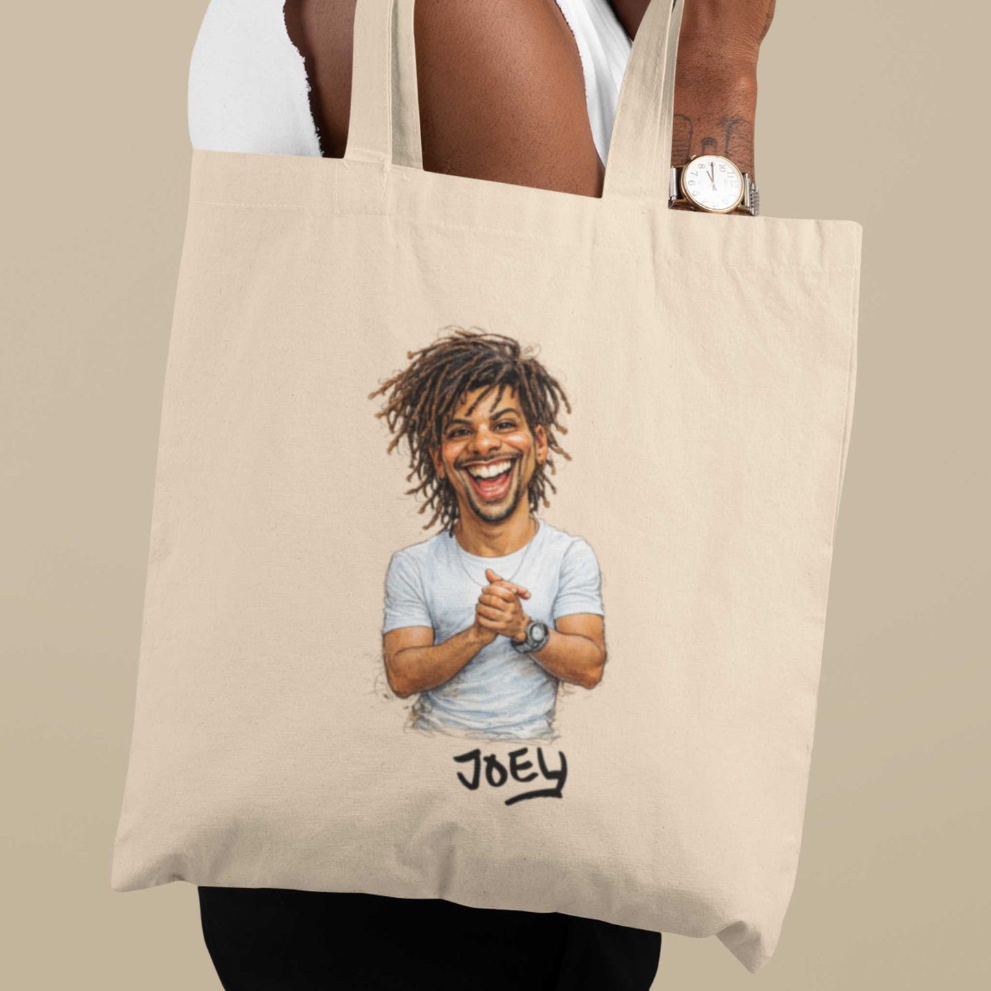 Beige katoenen totebag gedragen over de schouder, bedrukt met een gepersonaliseerde karikatuur van een lachende man met dreadlocks en de naam Joey onder de illustratie, gefotografeerd tegen een neutrale beige achtergrond.
