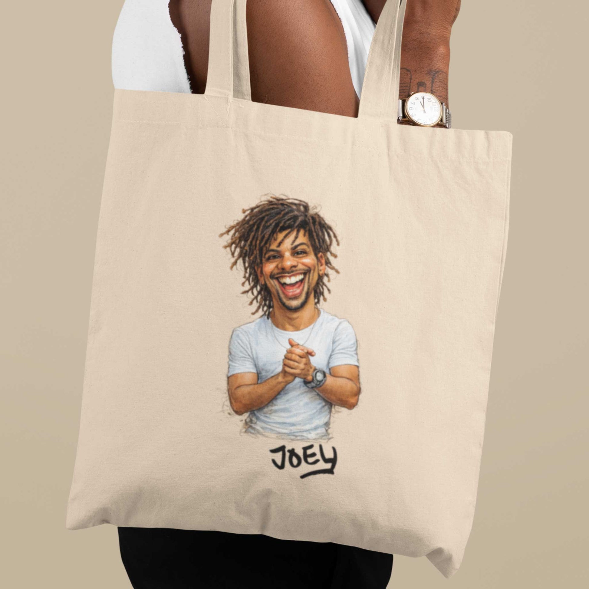 Beige katoenen totebag gedragen over de schouder, bedrukt met een gepersonaliseerde karikatuur van een lachende man met dreadlocks en de naam Joey onder de illustratie, gefotografeerd tegen een neutrale beige achtergrond.
